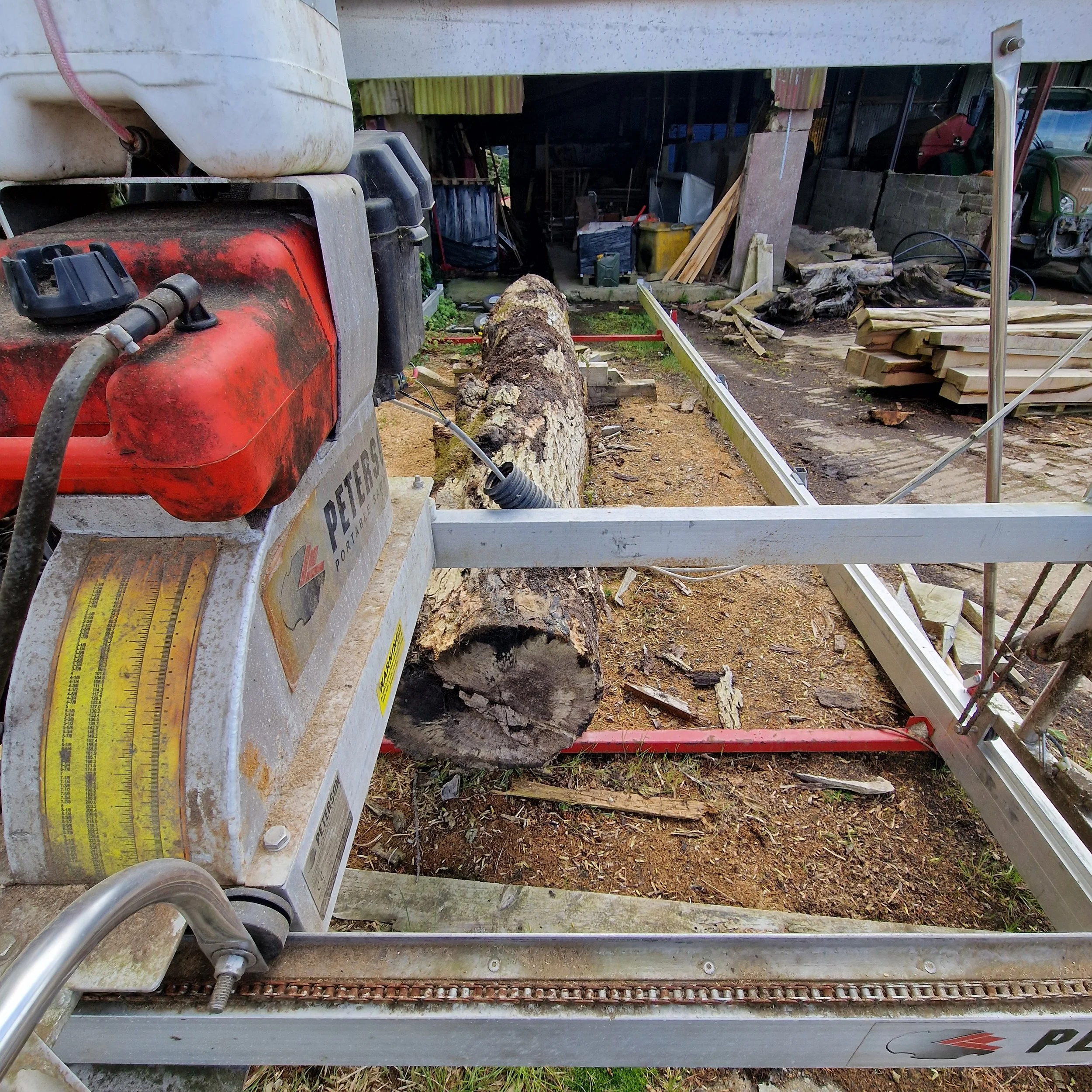A log mill setup with a chainsaw, grinding a large tree trunk into lumber in an outdoor workshop.