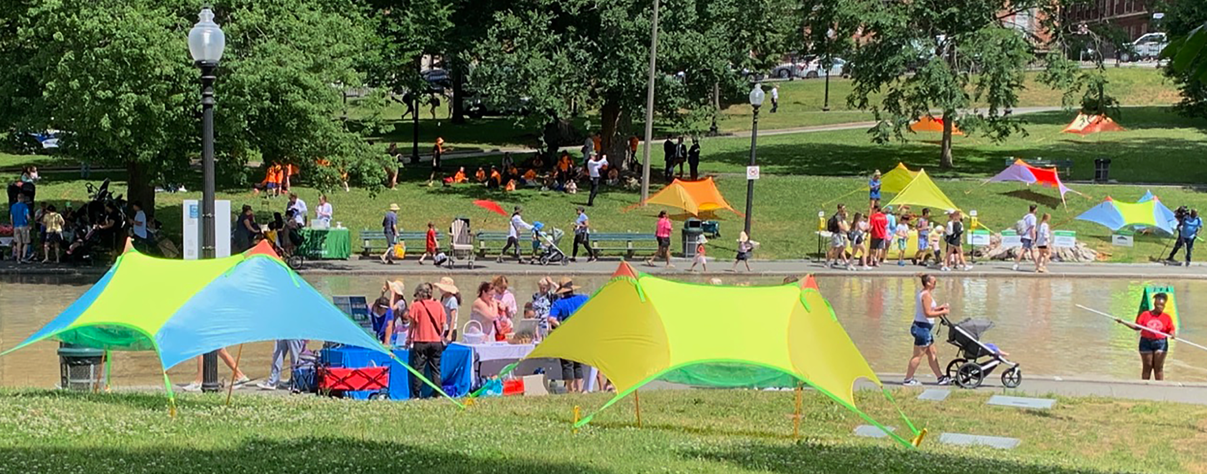 Crowds of people walking next to the Frog Pond at Boston Common with multiple colorful Zinnia sun canopy tents pitched on the grass.