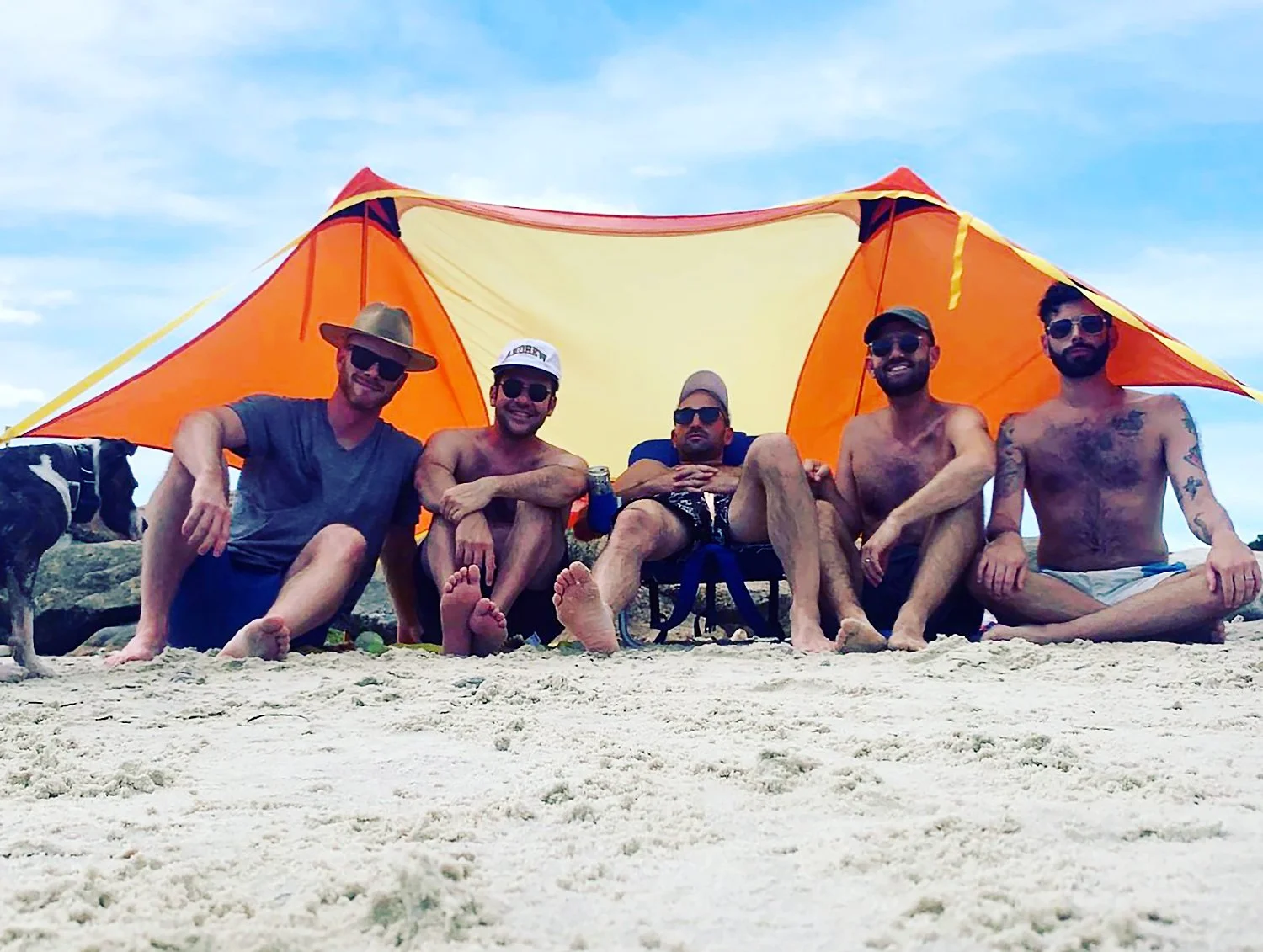 Five friends (bros) under a yellow and orange sun tent on a sandy beach, with a dog under the blue sky on a sunny day.