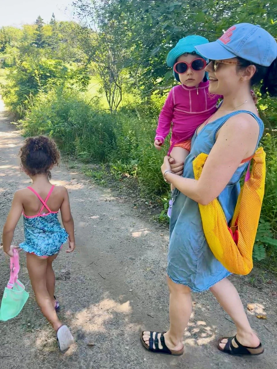 A woman carries her lightweight portable easy beach shade along with a child on her hip on the way to the beach in a lush green outdoor setting on a sunny day.