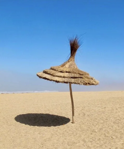 Classic thatched umbrella casting a circle of shade on the sand in the dessert under a clear blue sky.