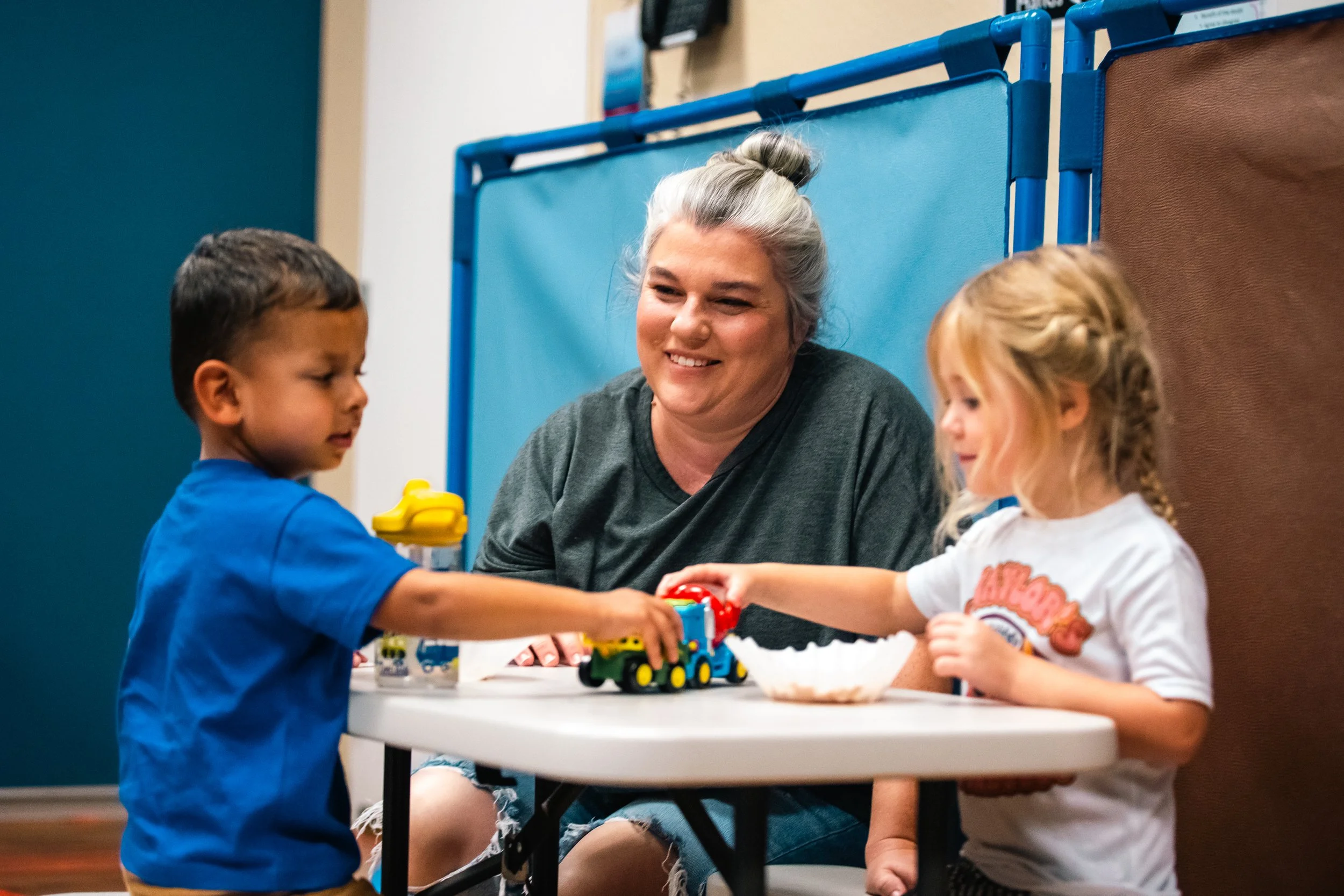 A woman with gray hair and two young children playing with colorful toy train set on a white table in a room with blue and brown walls.
