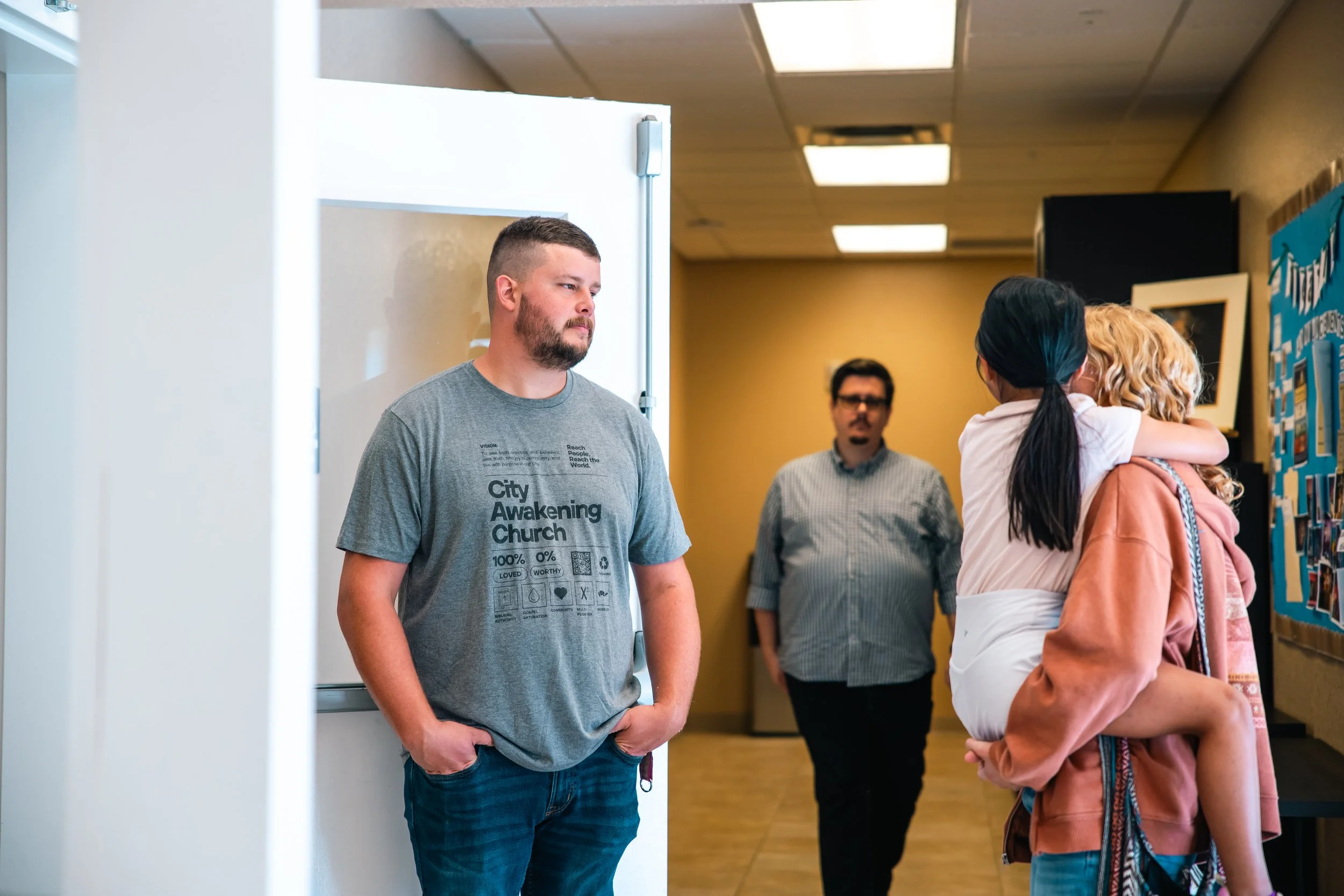 A man in a gray T-shirt and jeans stands near a white wall with his hands in his pockets, looking at a woman hugging a young girl. In the background, two other people are visible, including a man with glasses and a woman with dark hair.