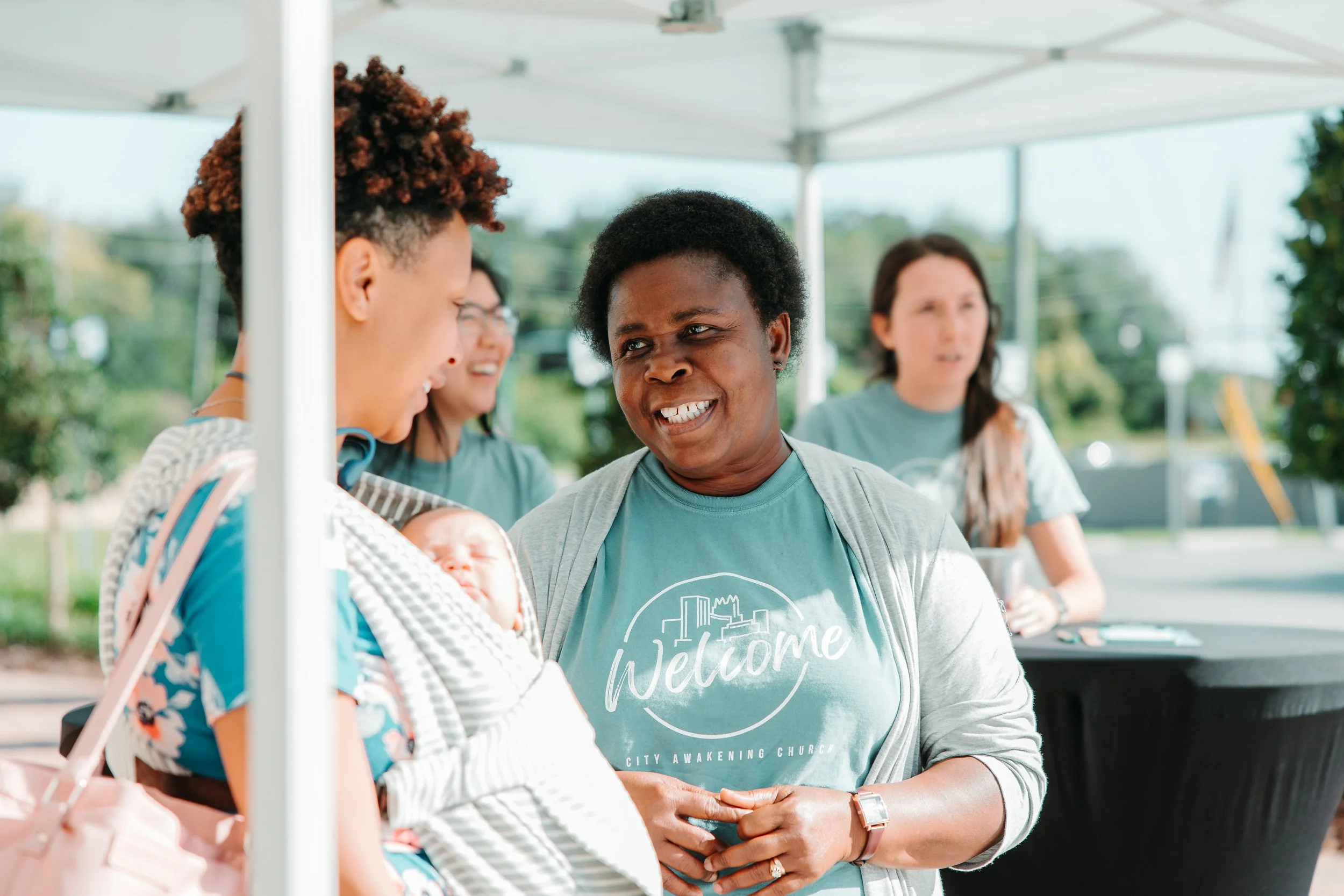 Group of people at outdoor event, with a woman holding a baby and talking to another woman wearing a gray shirt with 'Welcome' written on it.