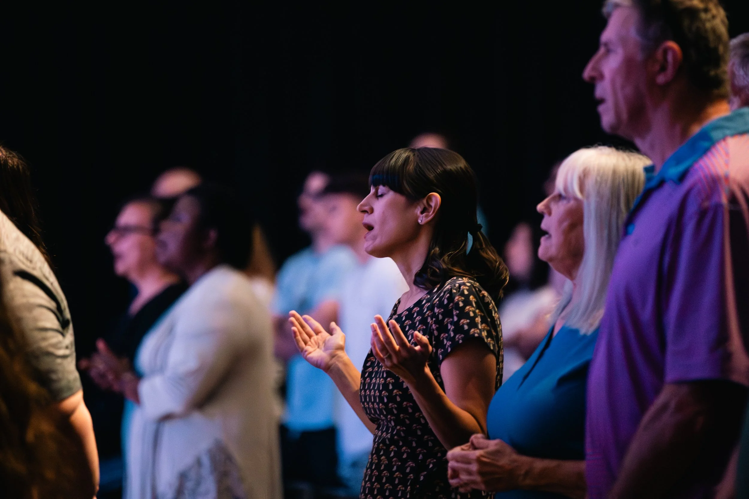 People standing with eyes closed and hands raised, participating in a prayer or worship session in a dimly lit environment.