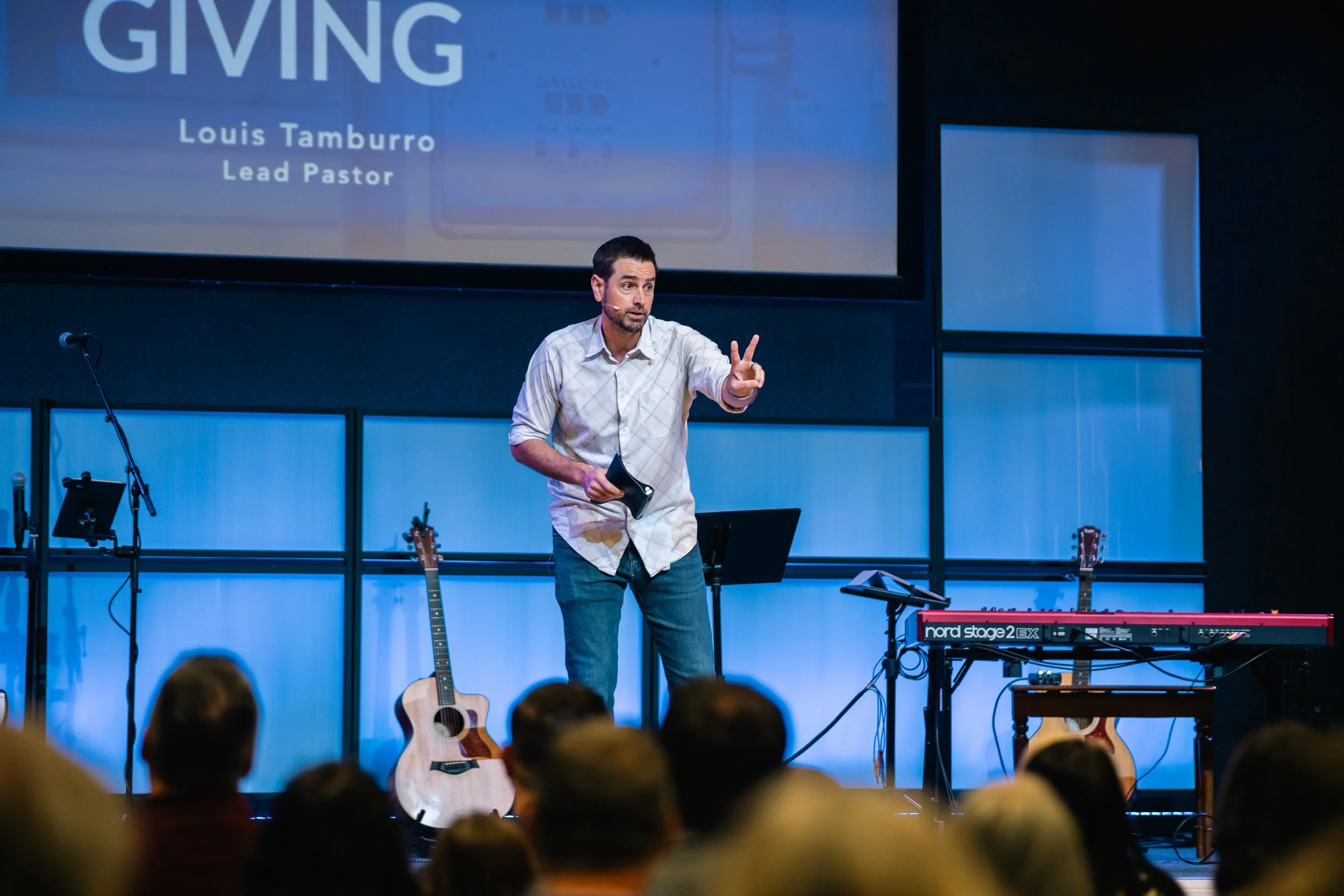 A man speaking on a stage during a church service or event, with a large screen behind him displaying 'GIVING' and his name, Louis Tamburro, Lead Pastor. The stage has musical instruments including guitars and a keyboard, and there are audience members visible in the foreground.