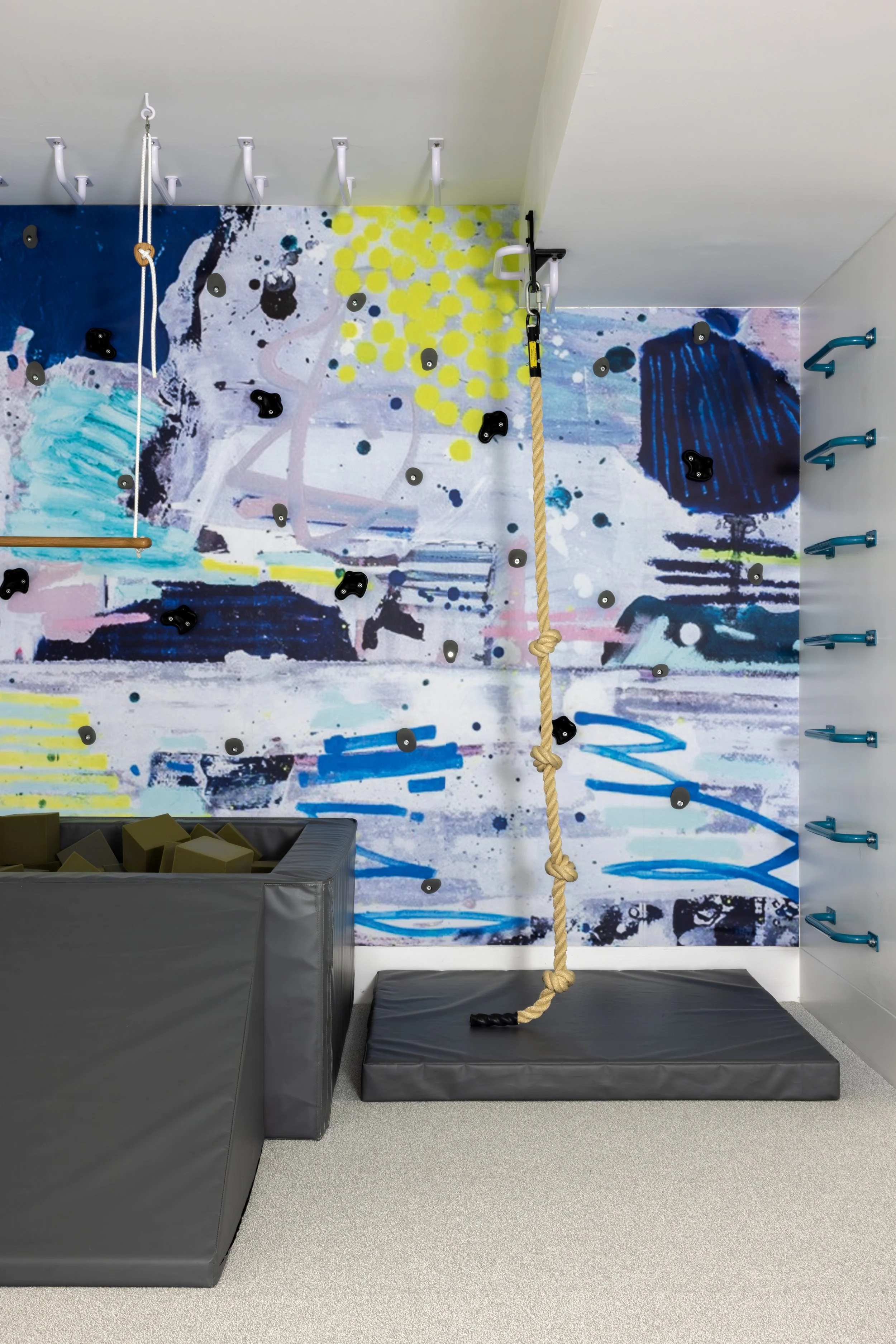 Indoor rock climbing gym with a colorful mural wall, a hanging climbing rope, padded floor, and foam blocks.