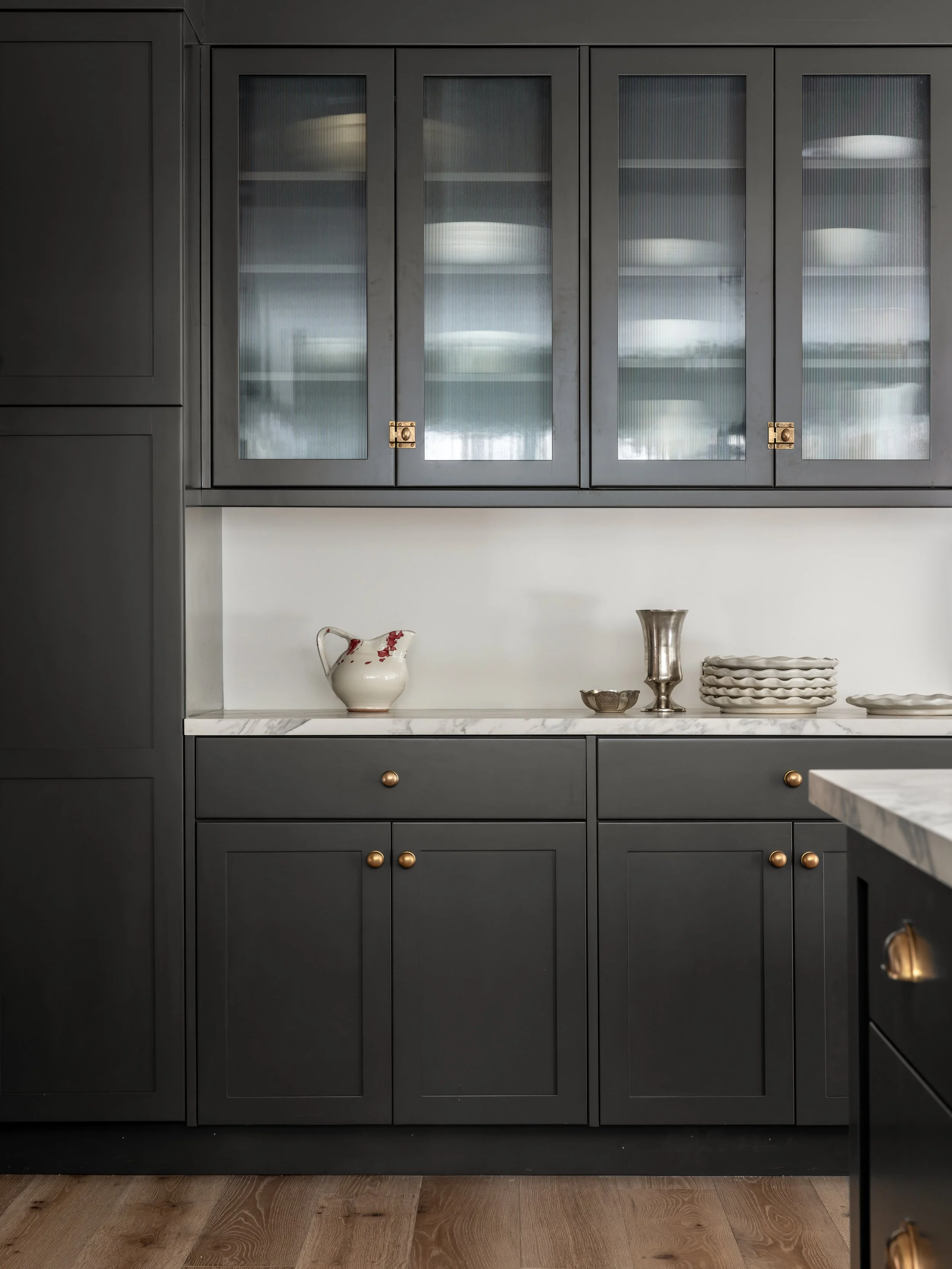 A kitchen with dark gray cabinets, a white marble countertop, and decorative dishes including a pitcher, bowls, and plates, with a wooden floor.