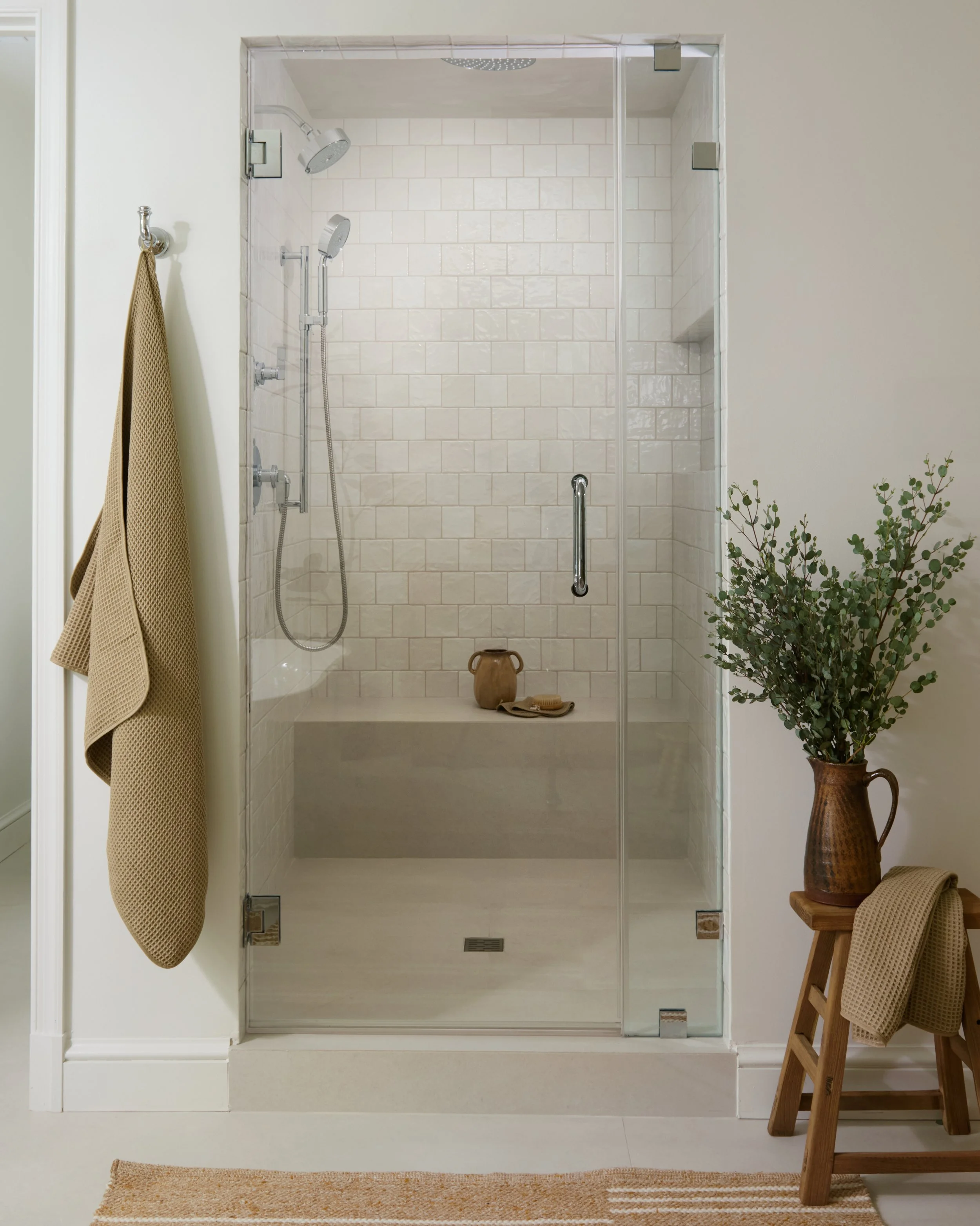 A modern walk-in shower with beige tiled walls, a glass door, and dual showerheads. To the left, a beige towel hangs on a silver hook. To the right, a wooden stool holds a brown ceramic pitcher, with a green leafy plant nearby. A beige bath mat is on