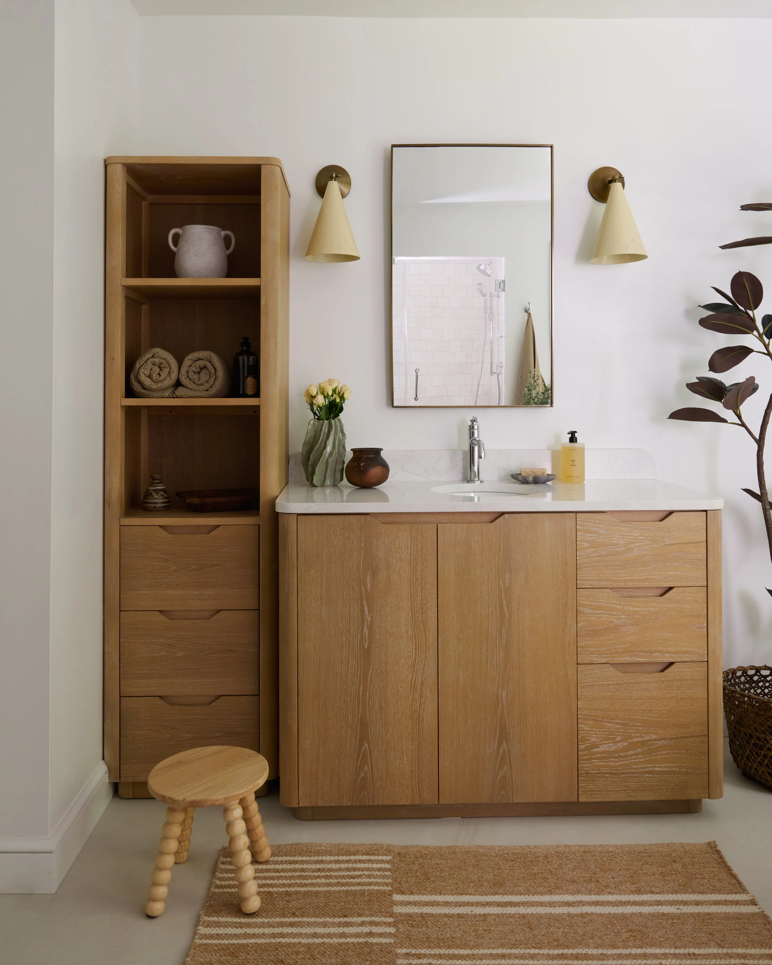 A bathroom vanity area with a wooden cabinet, a mirror, two wall-mounted lamps, and a small wooden stool in front of a striped rug. The countertop has a vase with flowers, a ceramic jar, a soap dispenser, and a small dish.