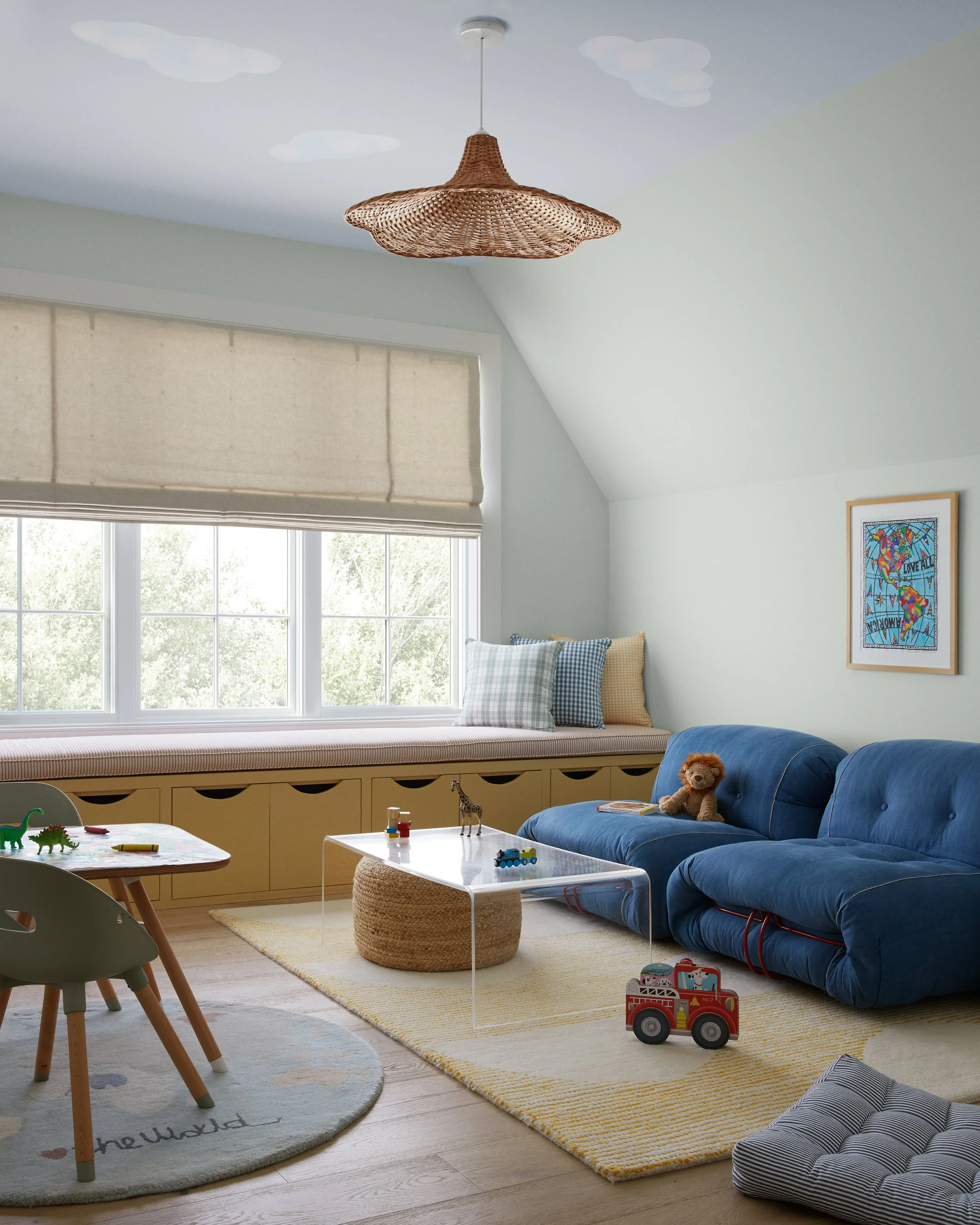 A cozy children's playroom with a large window, light-colored walls, and playful decor. There is a white window seat with checkered pillows, a yellow storage bench beneath it, and a kids' table with toys including dinosaurs and building blocks. A blu