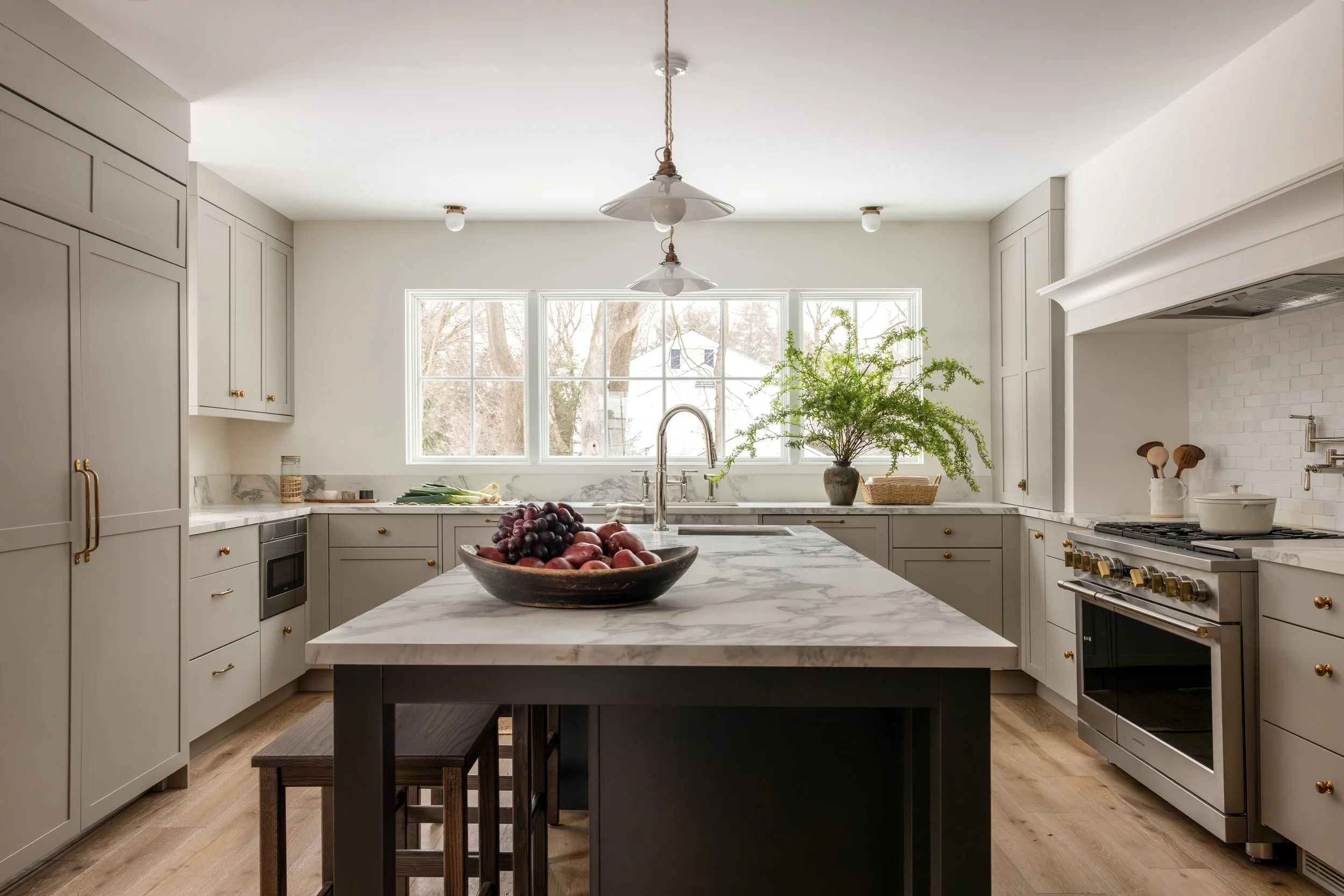 Bright kitchen with white cabinetry and a marble island, large window with trees outside, fruit bowl on island, potted plant near window, and modern appliances.