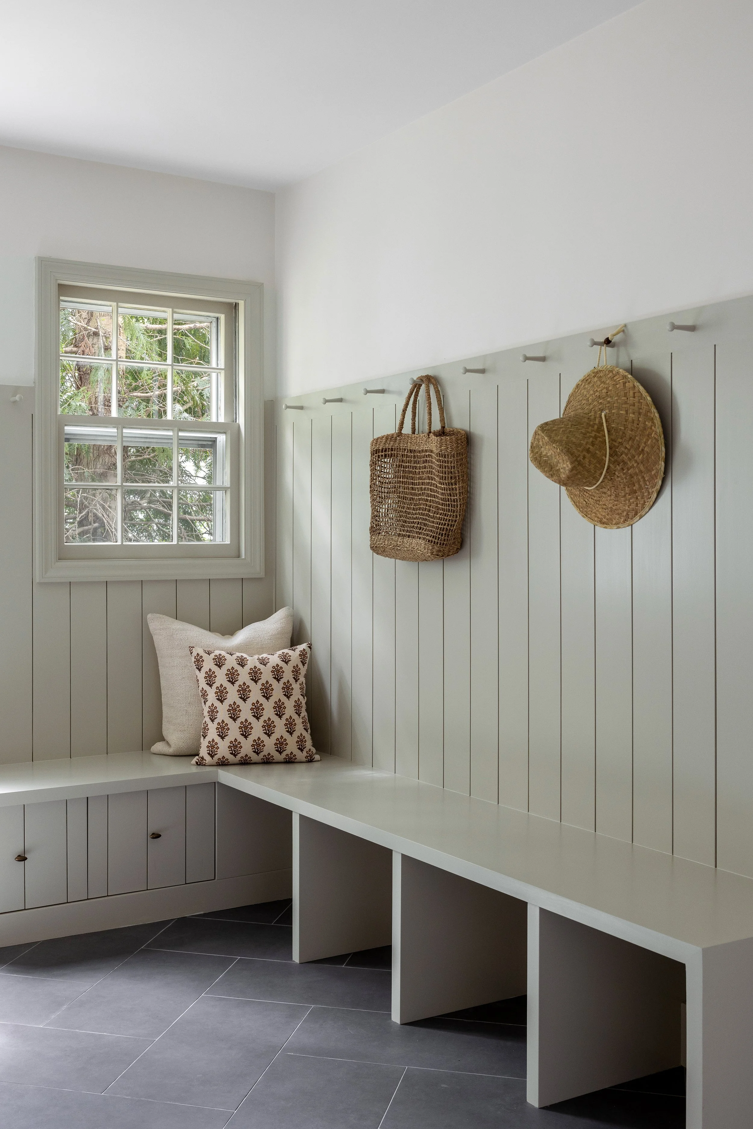 A corner of a room with white paneled walls, a window with greenery outside, baskets and a straw hat hanging on hooks, a built-in white bench with pillows, and dark gray tiled flooring.