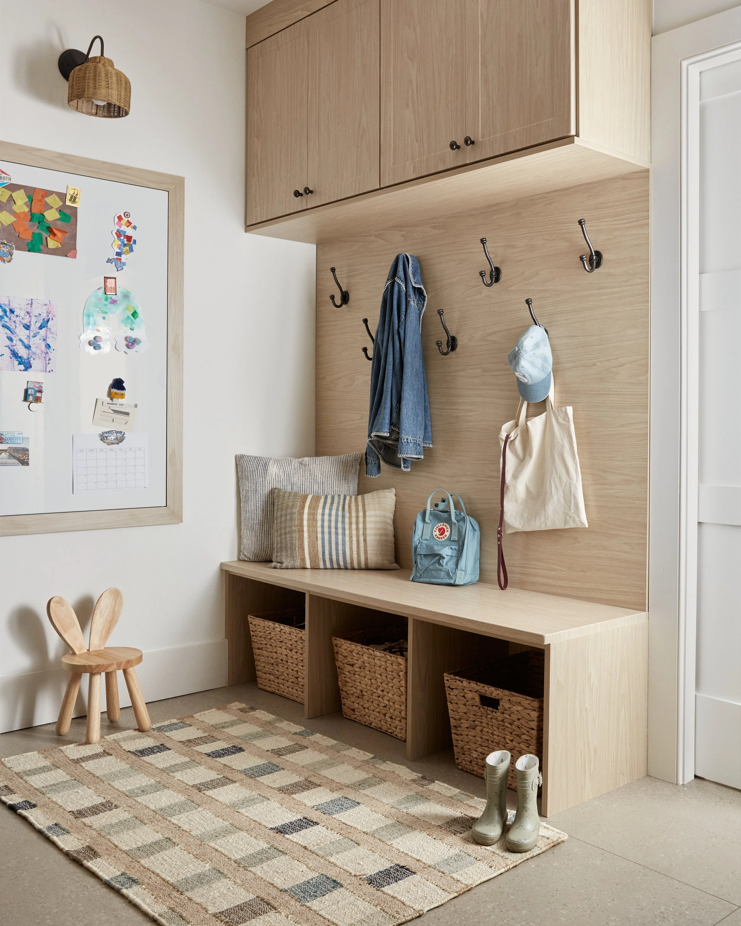Entryway with wooden bench, storage baskets, hanging hooks with jackets and bags, a pair of rain boots, a small wooden chair, a patterned rug, a whiteboard with colorful magnets, and overhead cabinet.