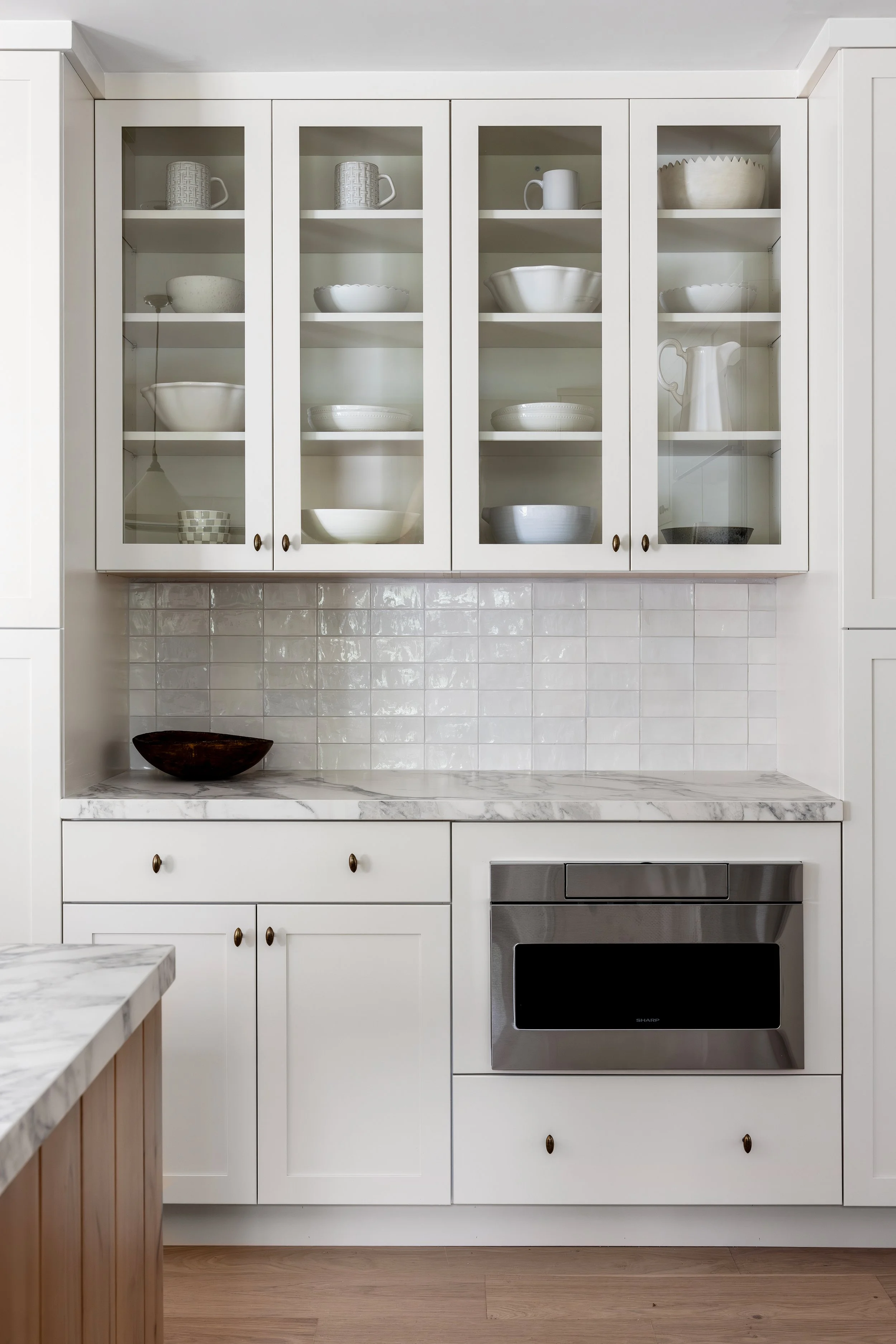Kitchen with white cabinets, glass-front upper cupboards displaying white dishes, bowls, cups, and a pitcher, and a built-in stainless steel microwave oven.
