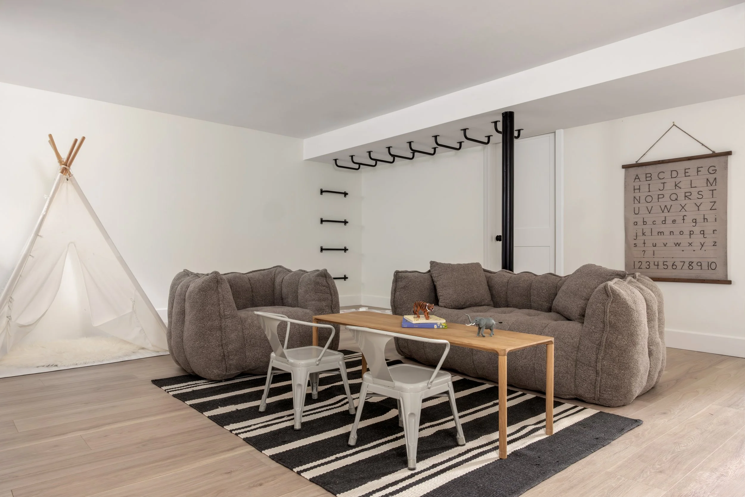 Living room with two large gray sofas, a wooden coffee table with toys on it, a small white children's chair, a black-and-white striped rug, a teepee tent, a wall hanging with the alphabet, and wall hooks for hanging items.