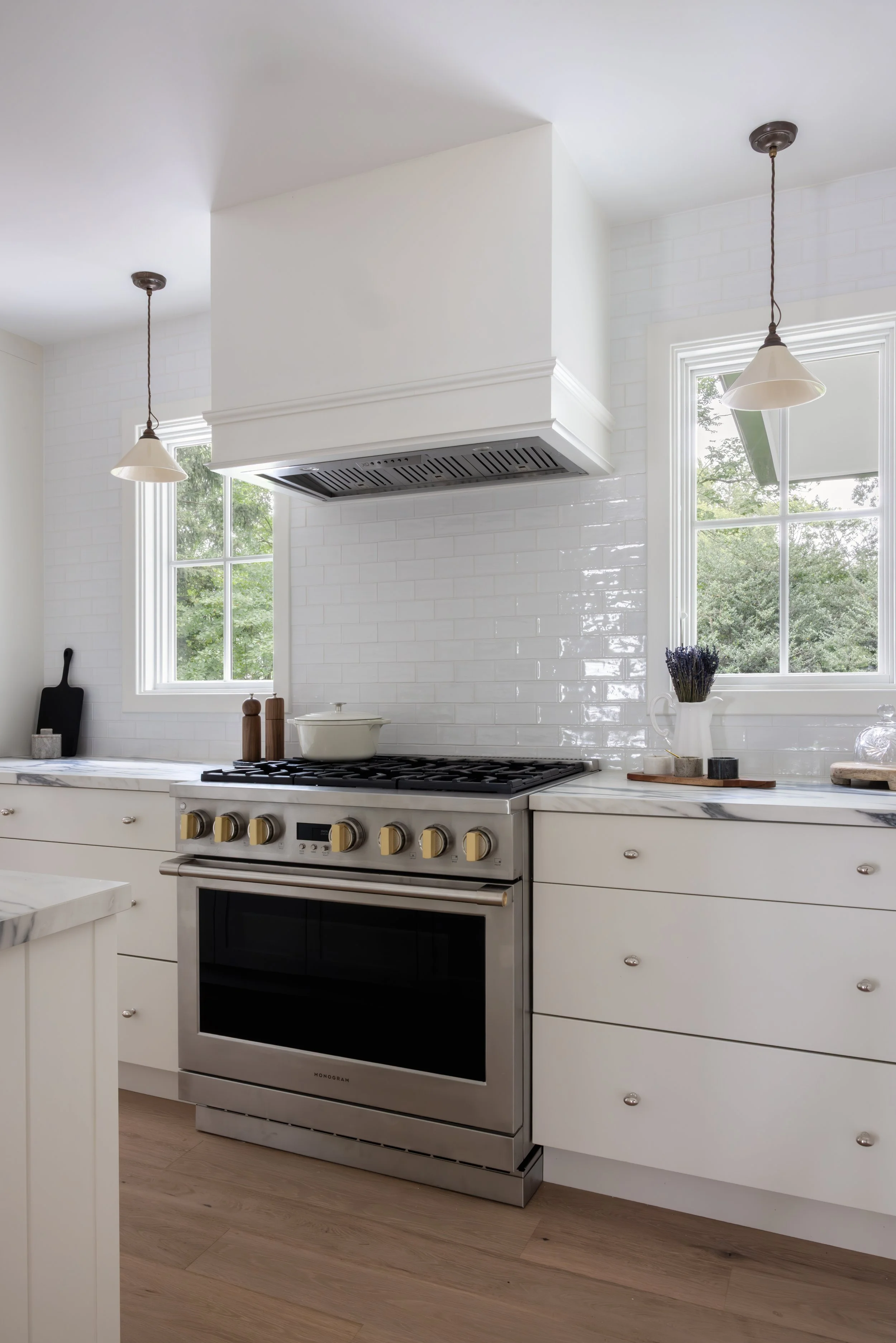 White kitchen with large windows, a stainless steel gas stove with black oven, white cabinets with silver knobs, marble countertop, and minimalistic decor including a vase of lavender and salt and pepper shakers.