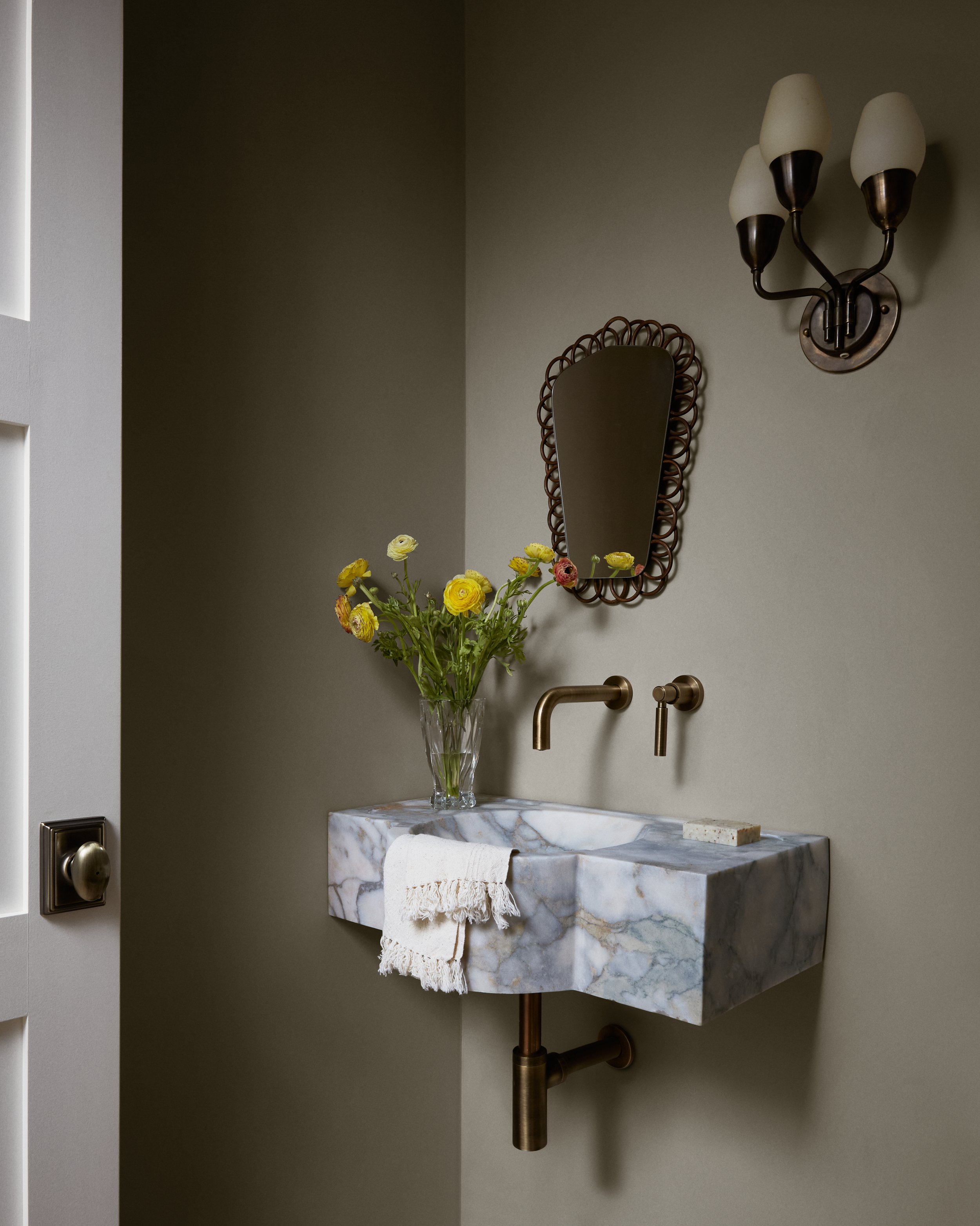 Bathroom sink with white marble countertop, a clear vase of yellow flowers, a mirror with a decorative frame, wall-mounted faucet, wall sconce with three shades, and a folded white towel.
