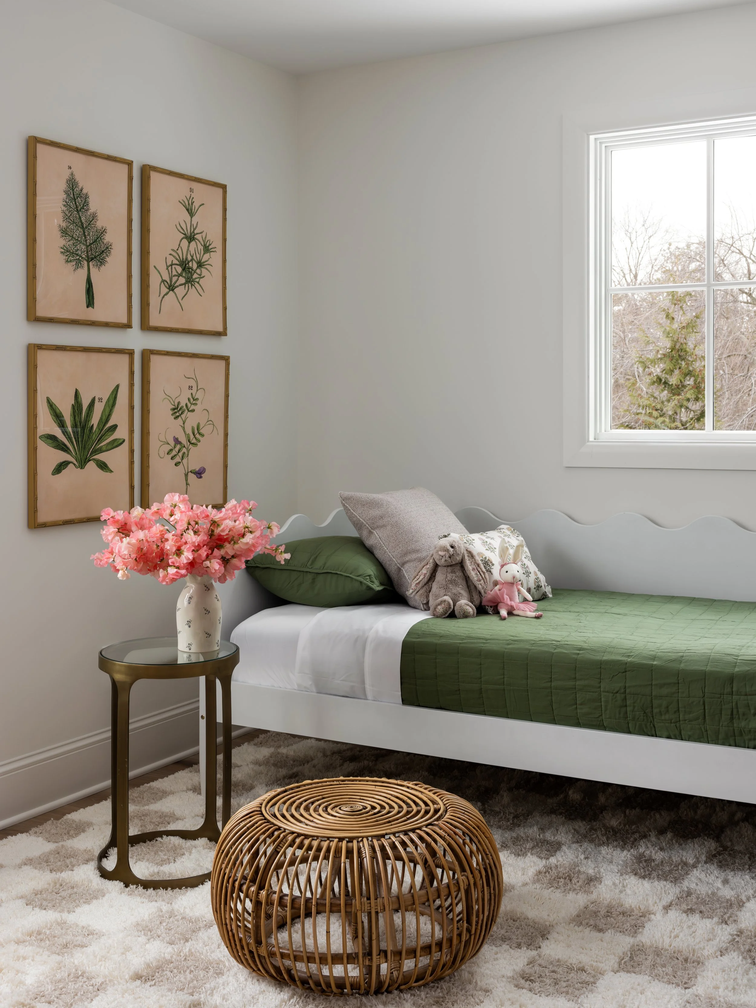 A bedroom with a white bed featuring a green quilt, set against a white wall with botanical artwork in bamboo frames, a side table with pink flowers in a white vase, plush toys on the bed, a window with a view of trees, and a rattan ottoman on a shag