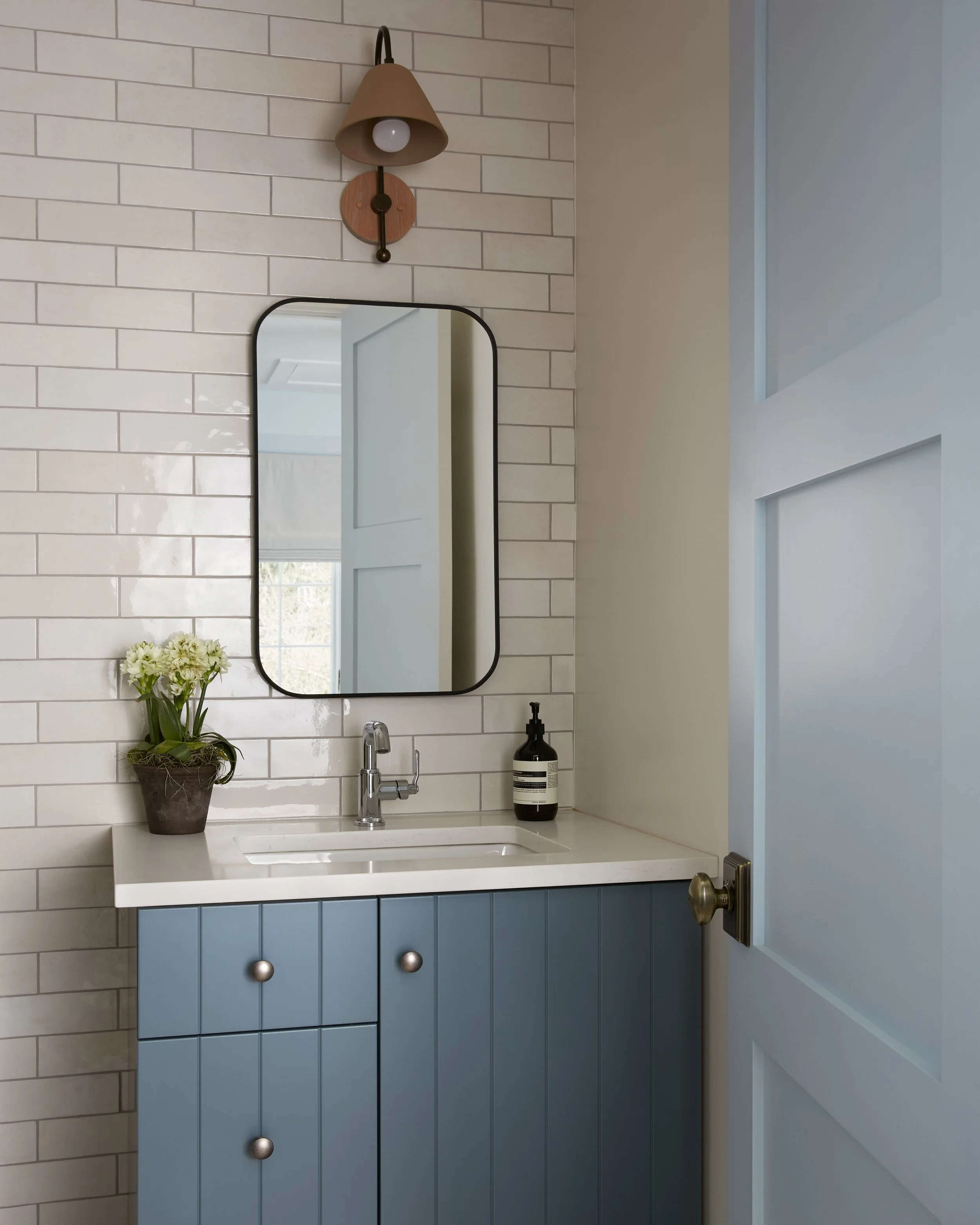 A bathroom with a rectangular mirror, a beige wall-mounted light fixture, a white countertop with a sink, a black soap dispenser, and a potted plant with white flowers. The door on the right is white with a gold doorknob.