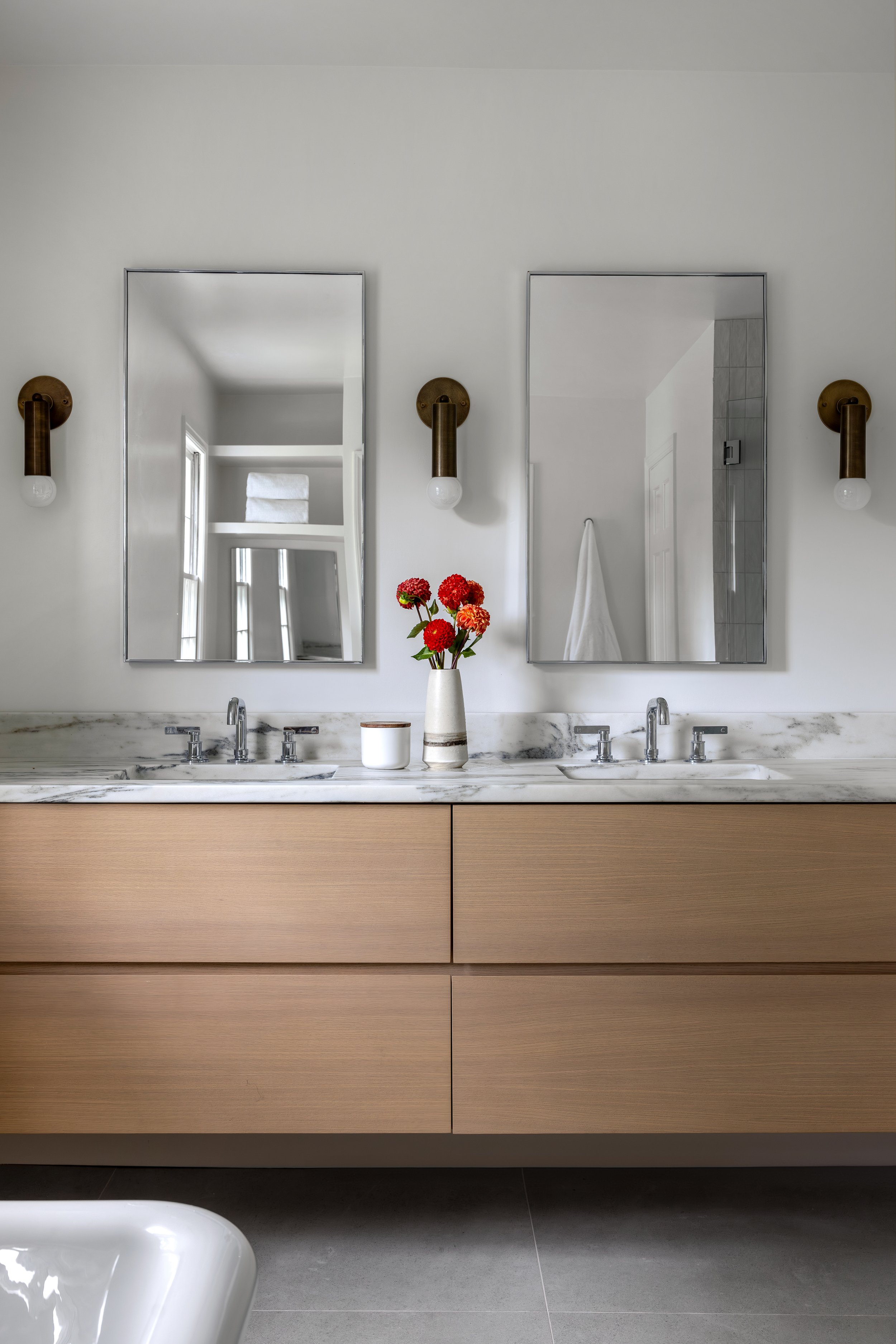Bathroom with two mirrors above a marble countertop with sinks, a vase of red and orange flowers, white towel on a shelf, wall-mounted light fixtures, and a white bathtub in the foreground.