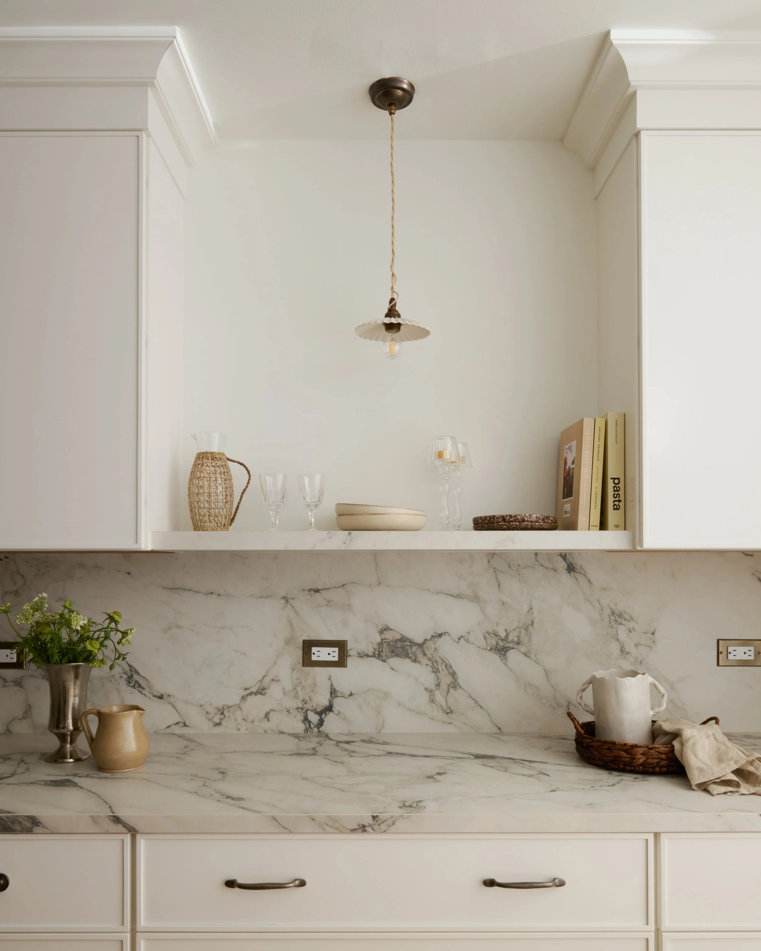A kitchen countertop made of white marble with dark veining. On the marble surface, there is a silver vase with green plants, a beige pitcher, and two beige pots. Behind the counter, there's a white open shelf with decorative items including glasswar