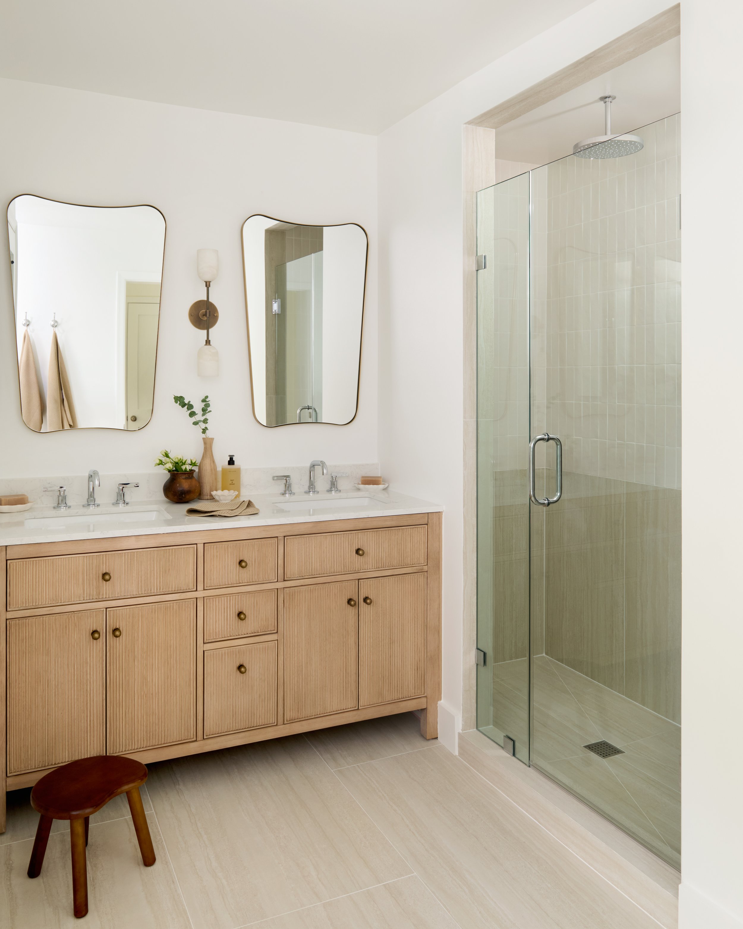 Bathroom with two mirrors, wooden vanity with soap and towels, small wooden stool, wall-mounted light fixture, and glass shower enclosure with beige tiles.