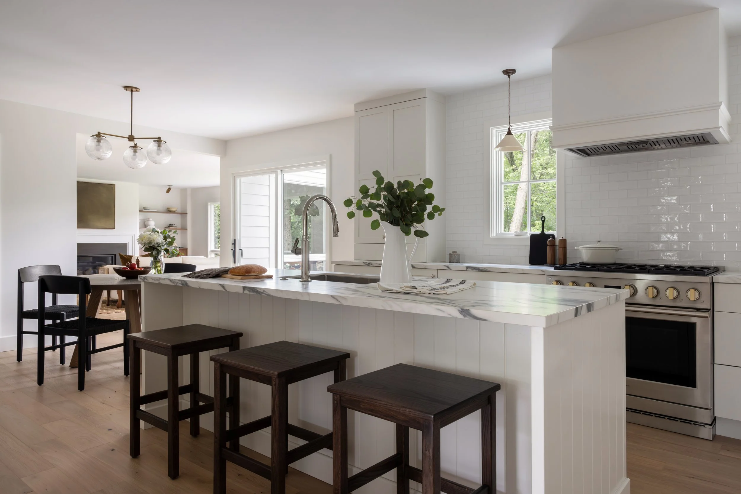 A modern kitchen with white cabinets, a marble island with a sink, black and wooden stools, and large windows with outdoor greenery.