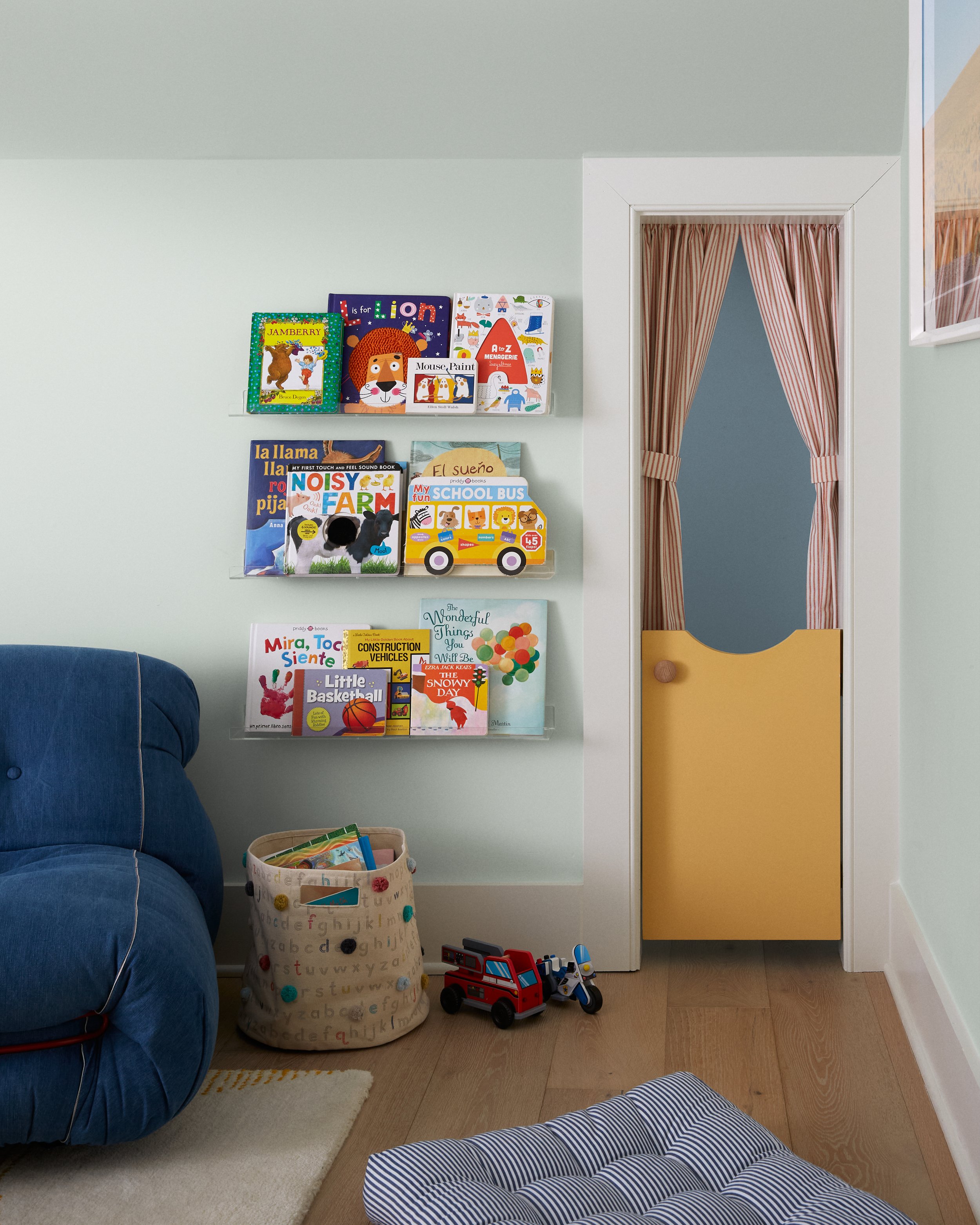 Children's books arranged on wall-mounted shelves next to a door with red and white striped curtains. A blue couch with a white rug and a basket filled with toys on the floor. Two toy trucks near a blue and white striped pillow.