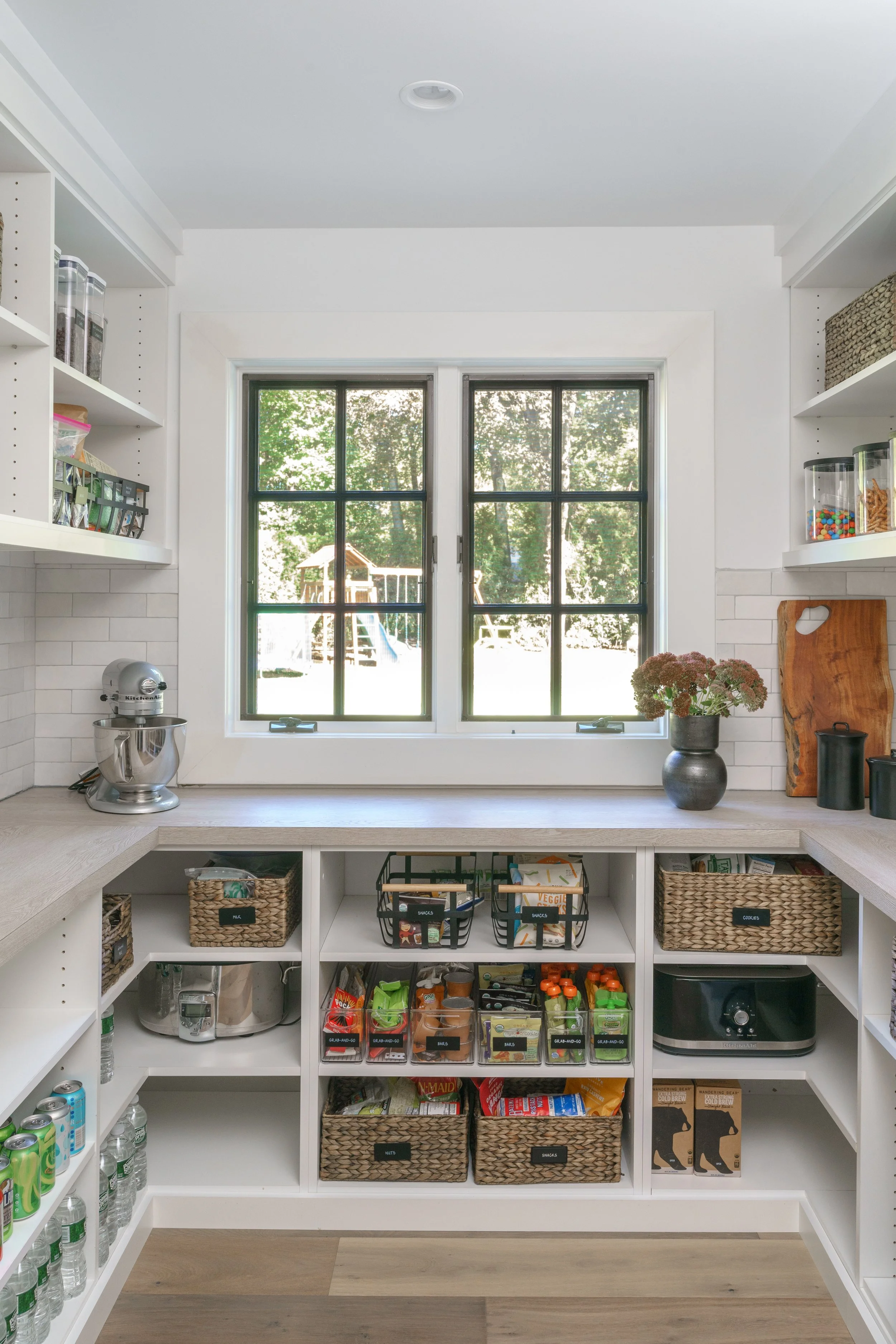 Kitchen pantry with organized shelves containing snacks, drinks, and kitchen appliances, with a window showing trees and a backyard outside.