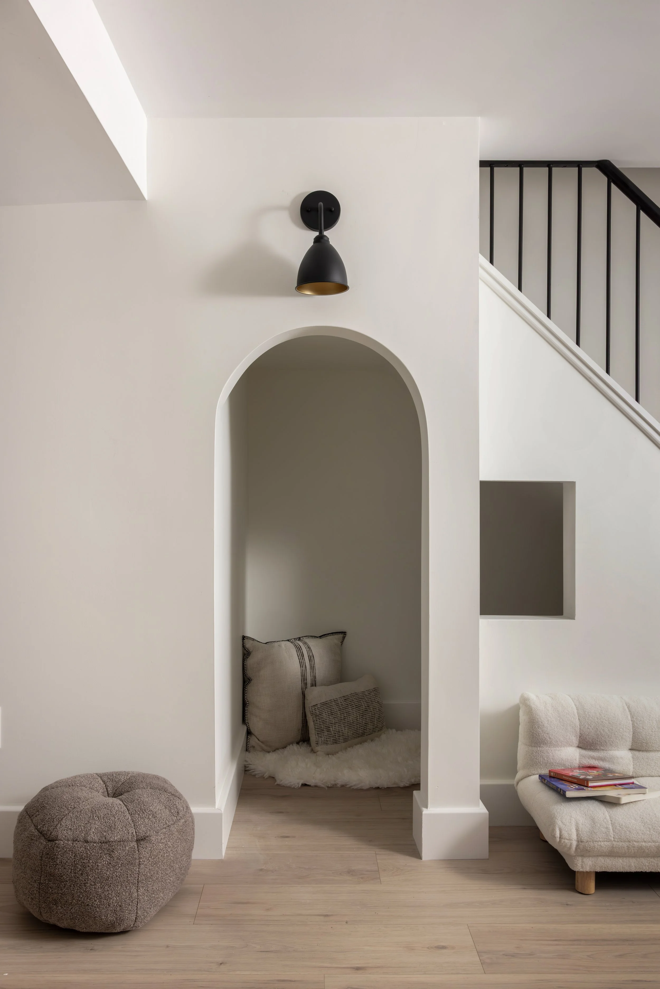 A cozy reading nook with a beige/gray armchair, books on the side, pillows in a small niche, and a fuzzy white rug on the floor. There is a black wall-mounted light fixture and staircase with black railing on the right.