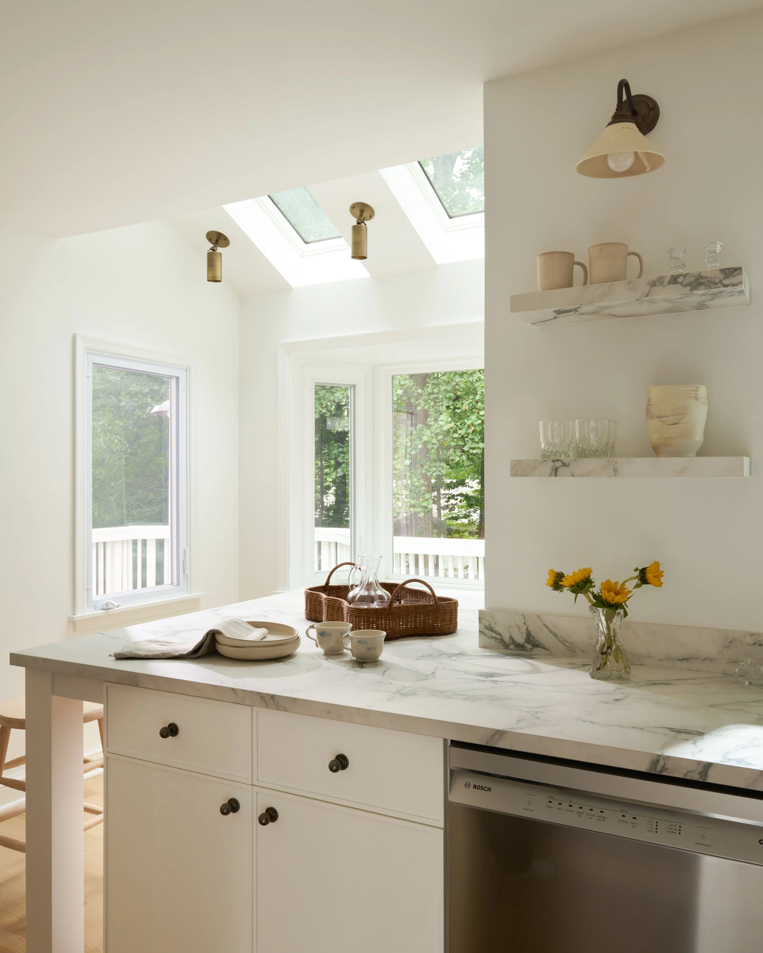 Kitchen with white cabinets, marble countertop, and a window with a view of green trees. Items on the counter include a glass pitcher, cups, a basket, a plate, and sunflowers. Two decorative shelves hold mugs, glasses, and a vase. Overhead lighting i