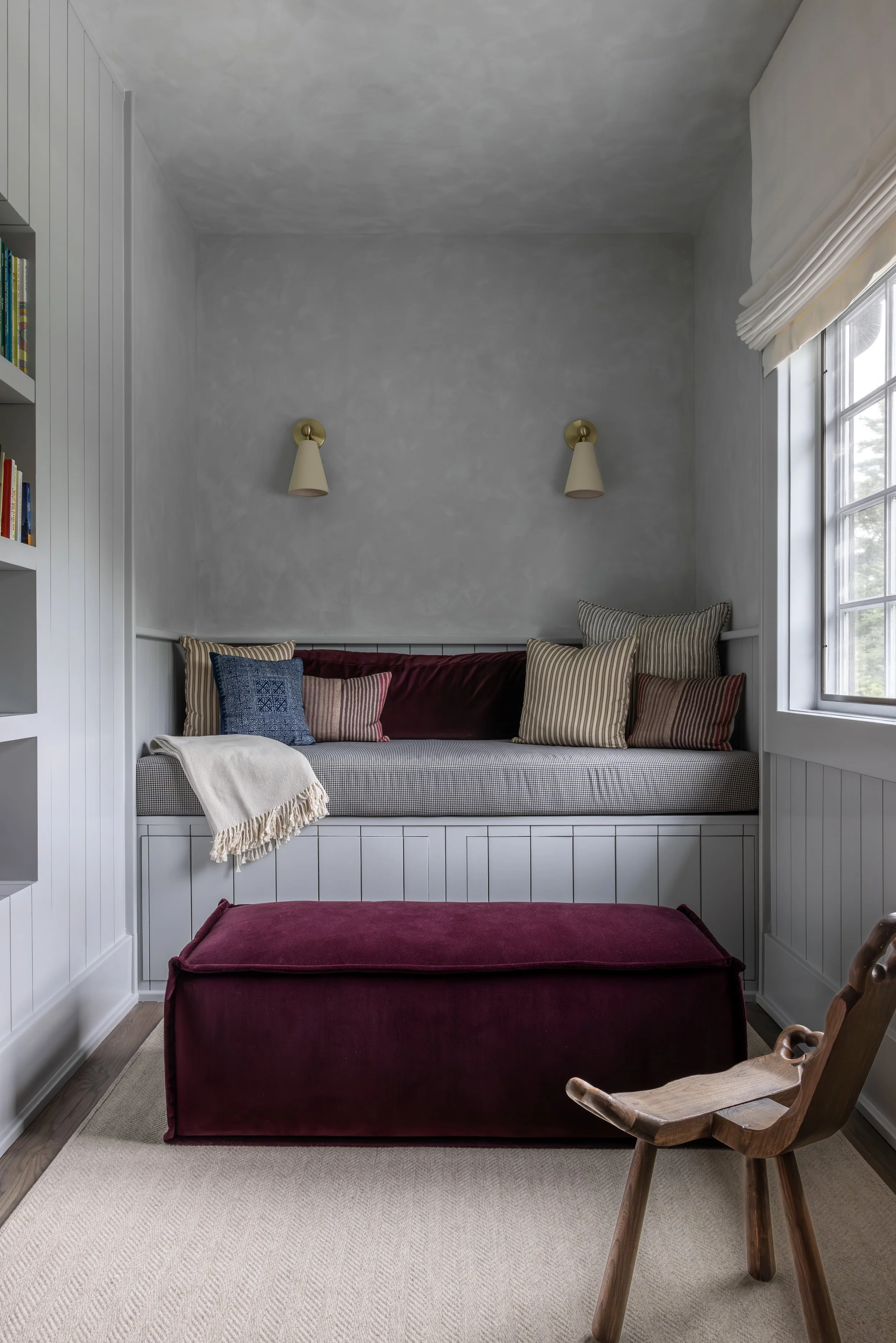 A cozy sitting area with a window, a gray cushioned window seat decorated with various striped and solid pillows, a blanket, and a red velvet ottoman in front. There are two wall-mounted lamps above the window seat, and part of a wooden rocking chair