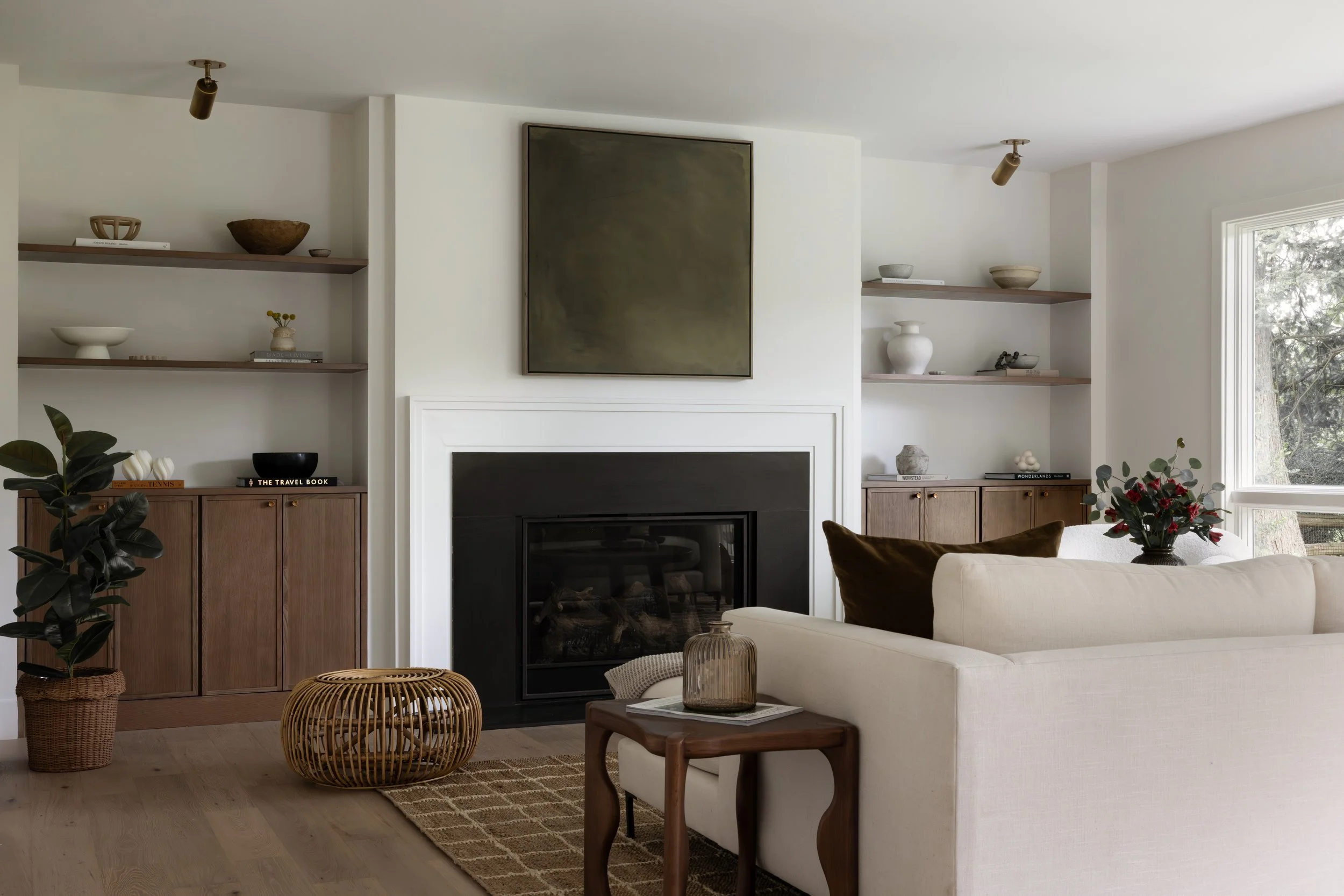 A modern living room with a white fireplace, wooden shelves with decorative bowls and books, a cream sofa with brown pillows, a wooden side table with a glass vase, plants, and natural light coming through a window.