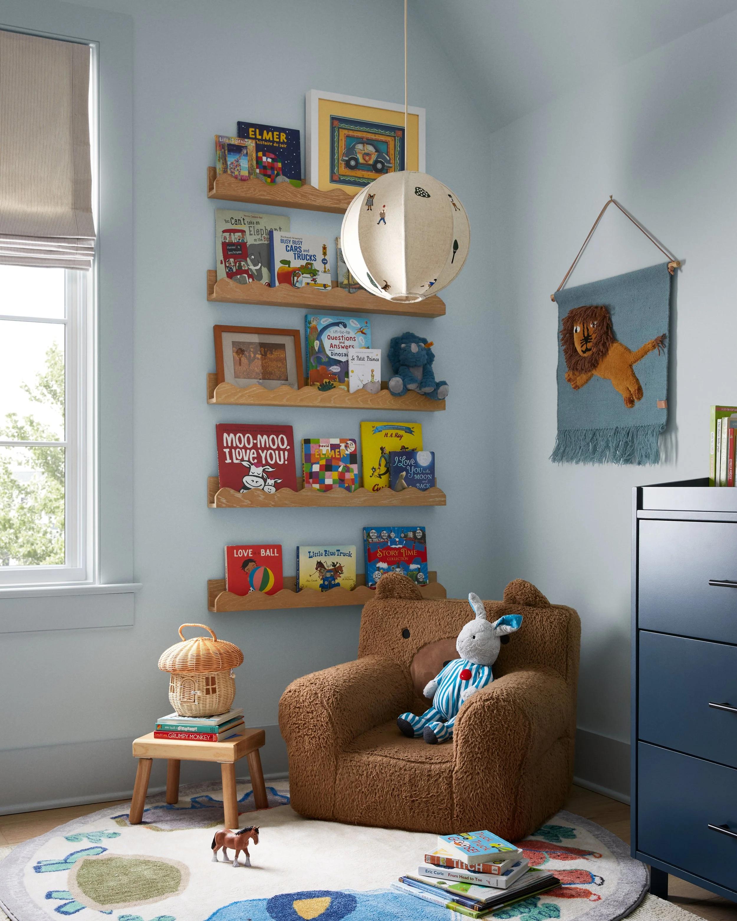 A cozy children's reading nook with a plush brown armchair, stuffed animals including a dog and a rabbit, and a stack of books on the floor. There are wooden shelves filled with children's books on the wall and a small side table with a woven lamp. A