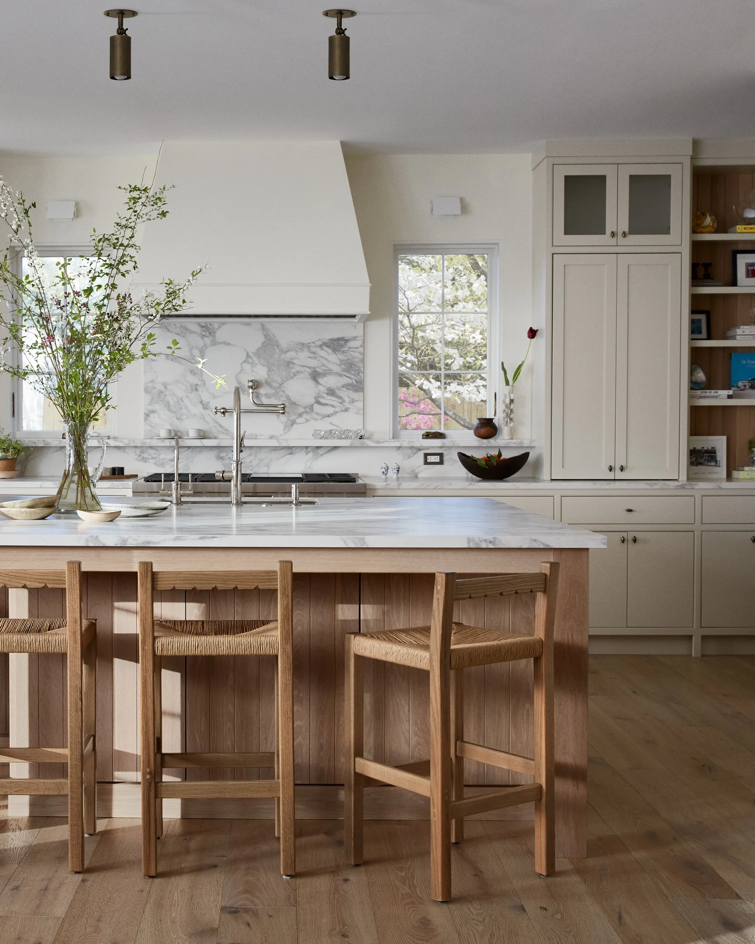 A kitchen with a marble backsplash, a large white kitchen island with a marble countertop, and four wooden chairs with woven seats. There are two windows, one on each side, with outside trees visible. The kitchen has built-in cabinets and open shelve
