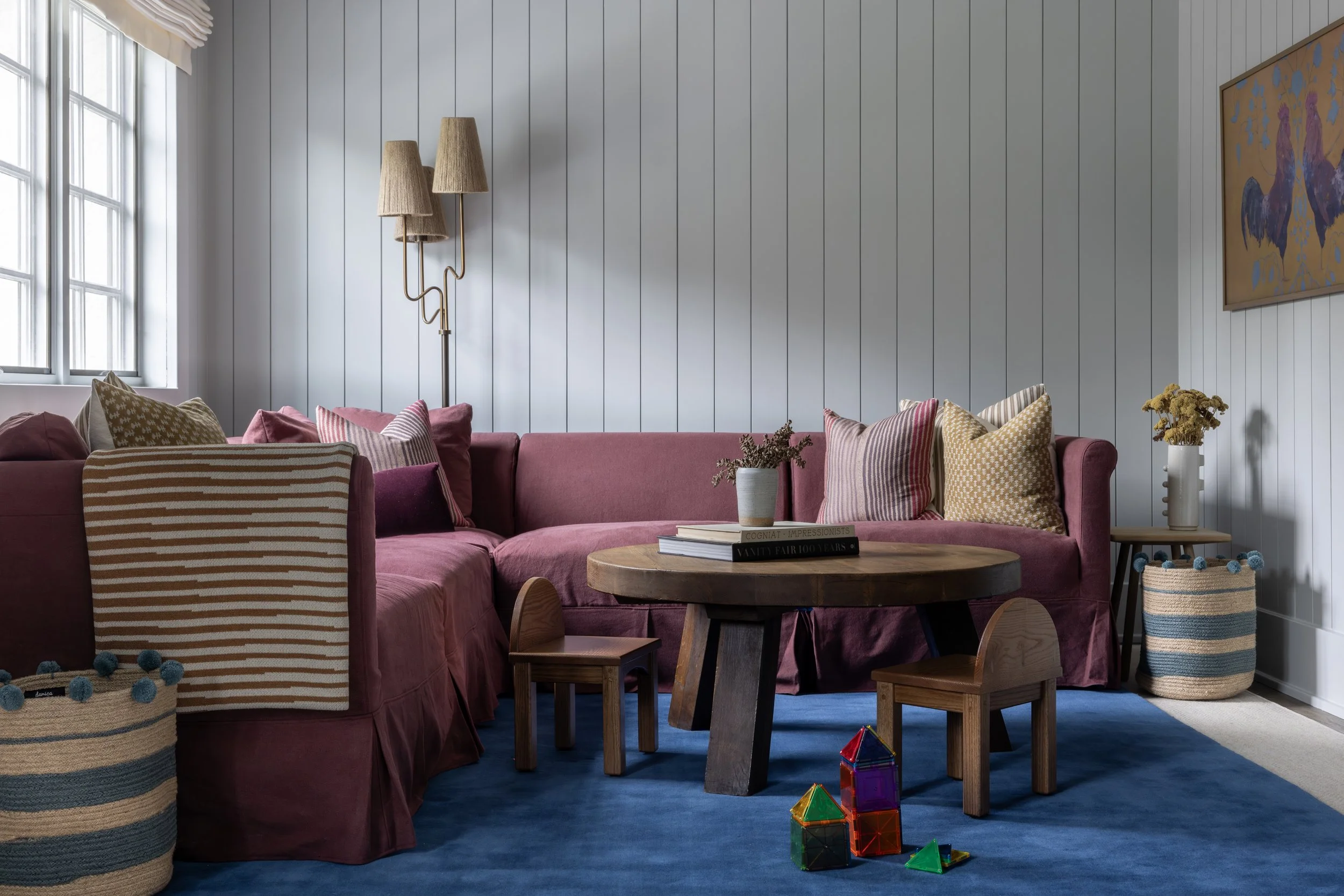 Living room with a large pink sofa, decorated with patterned pillows, against a white paneled wall. A wooden coffee table holds a stack of books, with playful building blocks on the blue carpeted floor. To the right, a tall basket contains yellow flo