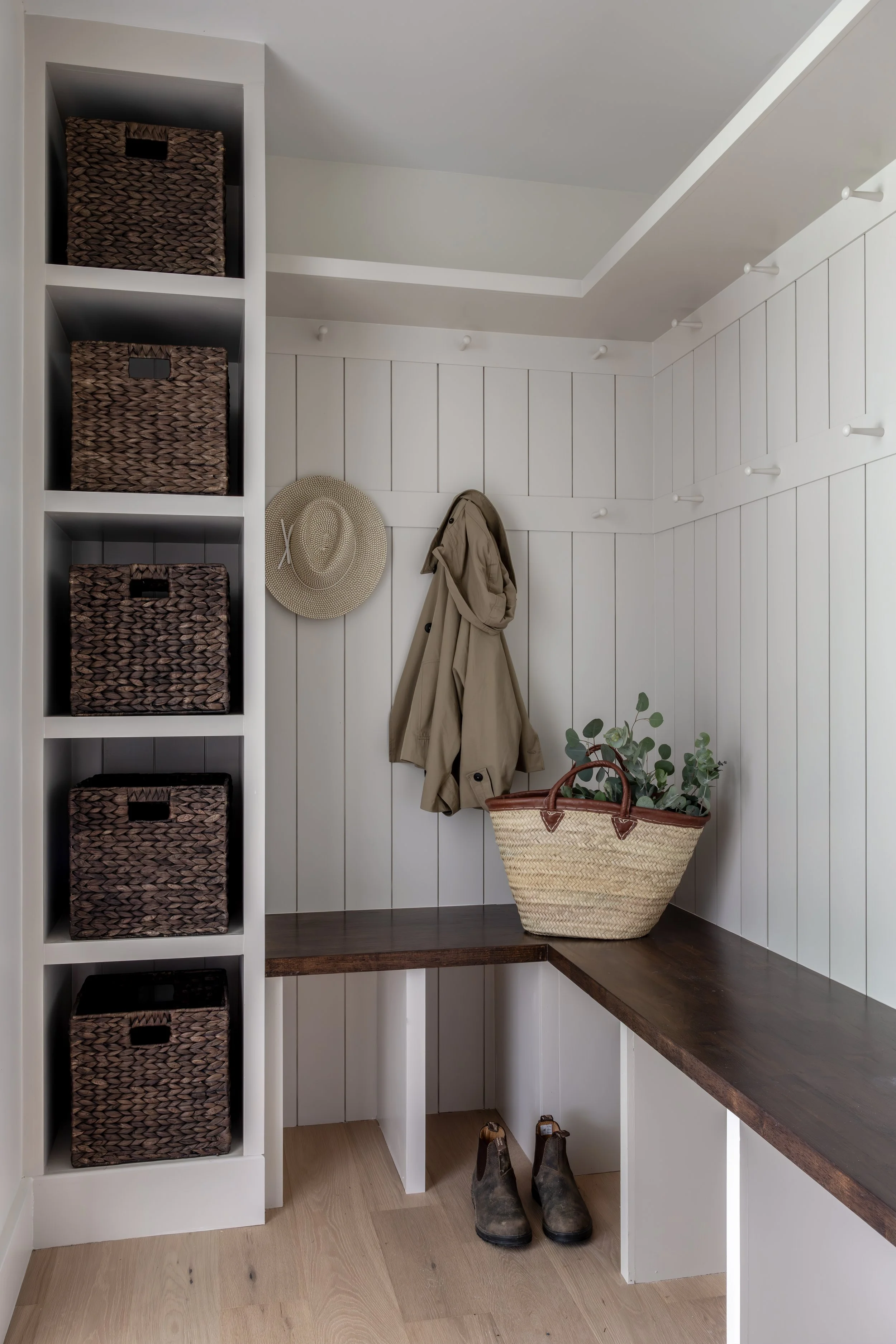 A small entryway with white paneled walls and a wooden floor. There are four woven baskets on a white corner shelf, a straw hat hanging on the wall, a beige coat also hanging, a basket with plants on a wooden shelf, and a pair of brown boots on the f