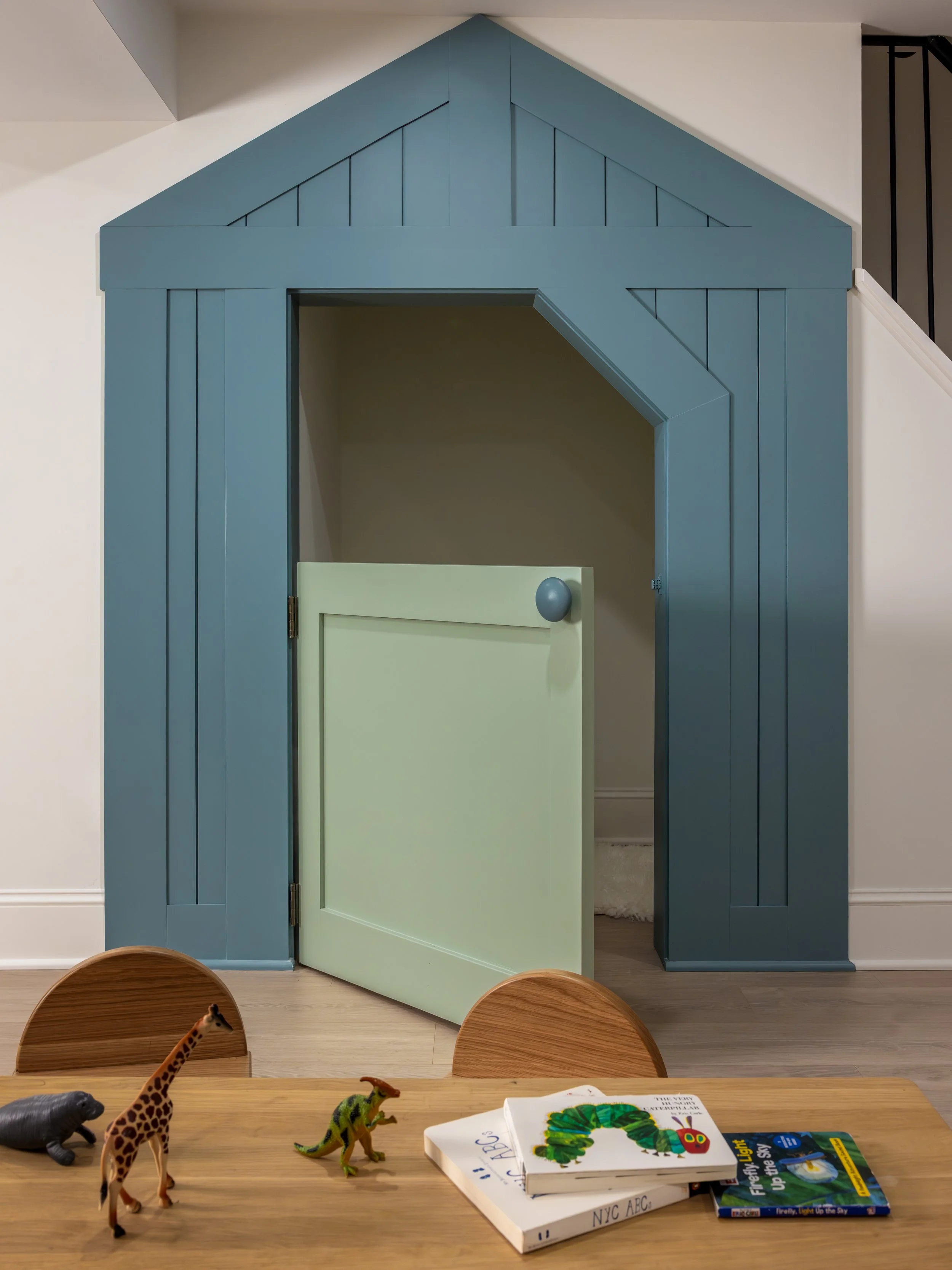View of a blue wooden playhouse or structure with a partially open small door, located in a room with a white wall and wood flooring. In the foreground, a wooden table has children's books and plastic animal toys including a giraffe, a dinosaur, and 