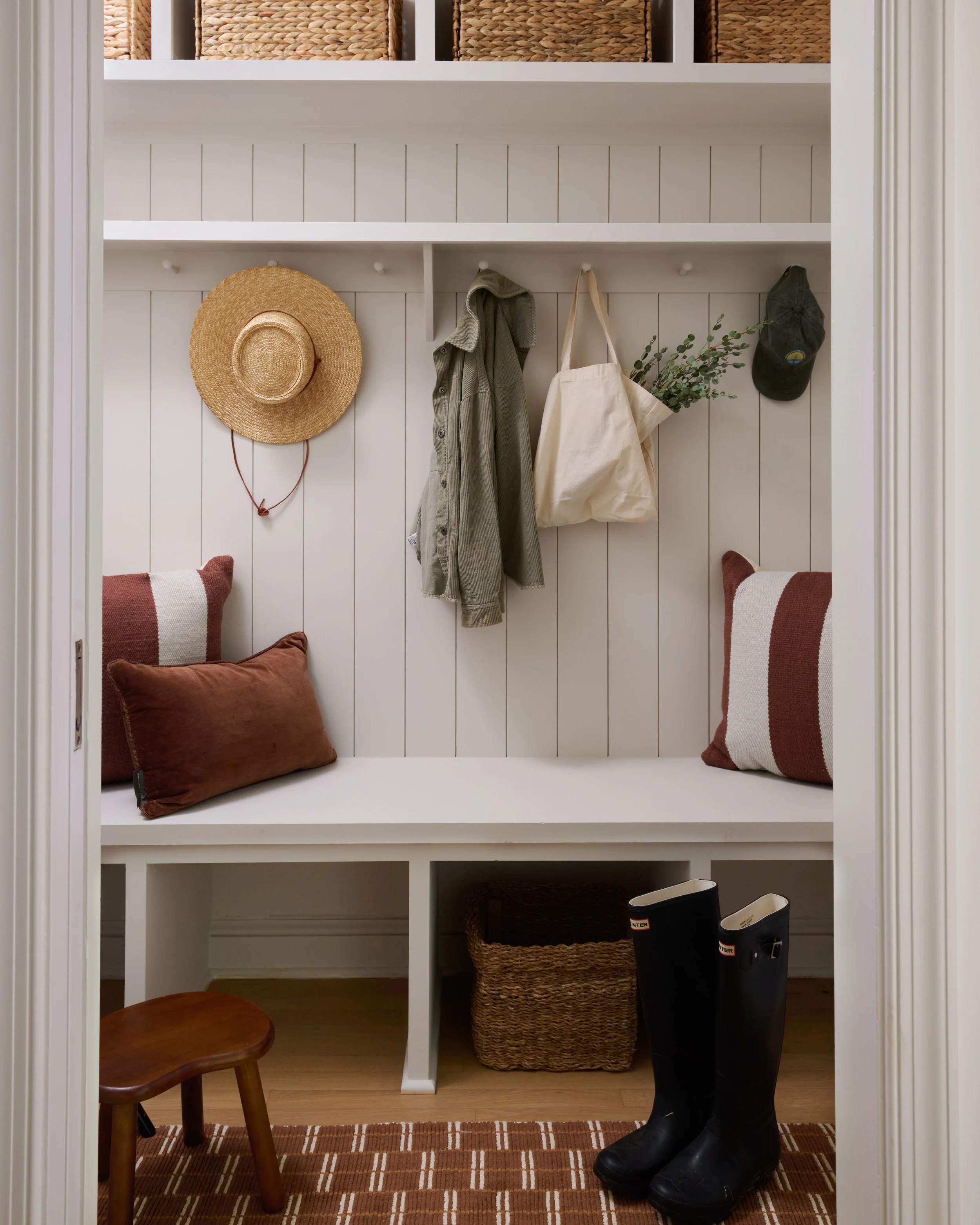 A white built-in closet with shelves and hooks, holding a straw hat, a green jacket, a beige tote bag with greenery, and a black cap. The bottom shelf contains a brown pillow, a woven basket, and black rubber boots on a brown striped rug.