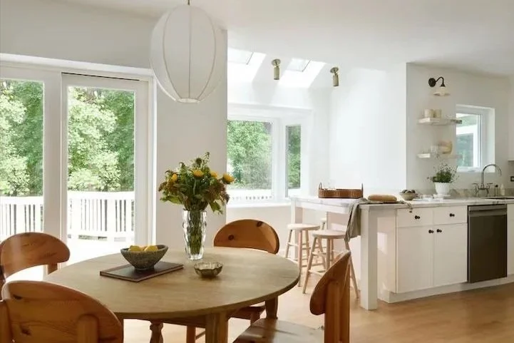 Bright, airy kitchen and dining area with white walls, wooden dining table with four chairs, vase with yellow flowers, large sliding glass doors leading outside, small breakfast bar with two stools, white cabinets, and a window above the sink.
