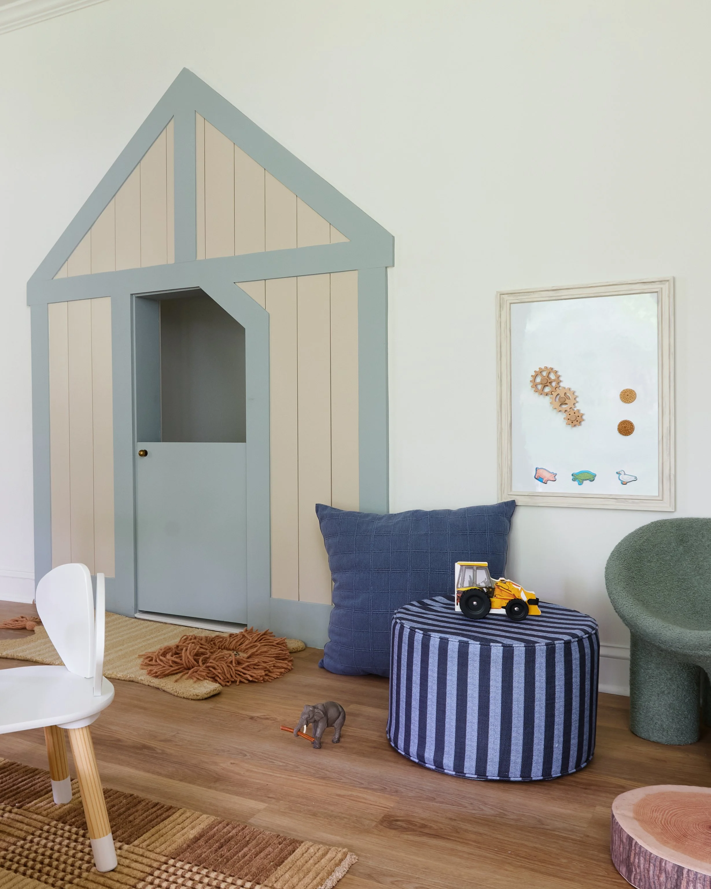 Children's playroom with a large blue and white wooden playhouse, a striped blue and white ottoman with a toy construction vehicle on top, a green textured chair, and a small white chair. There are decorative gears and animal cutouts on the wall.