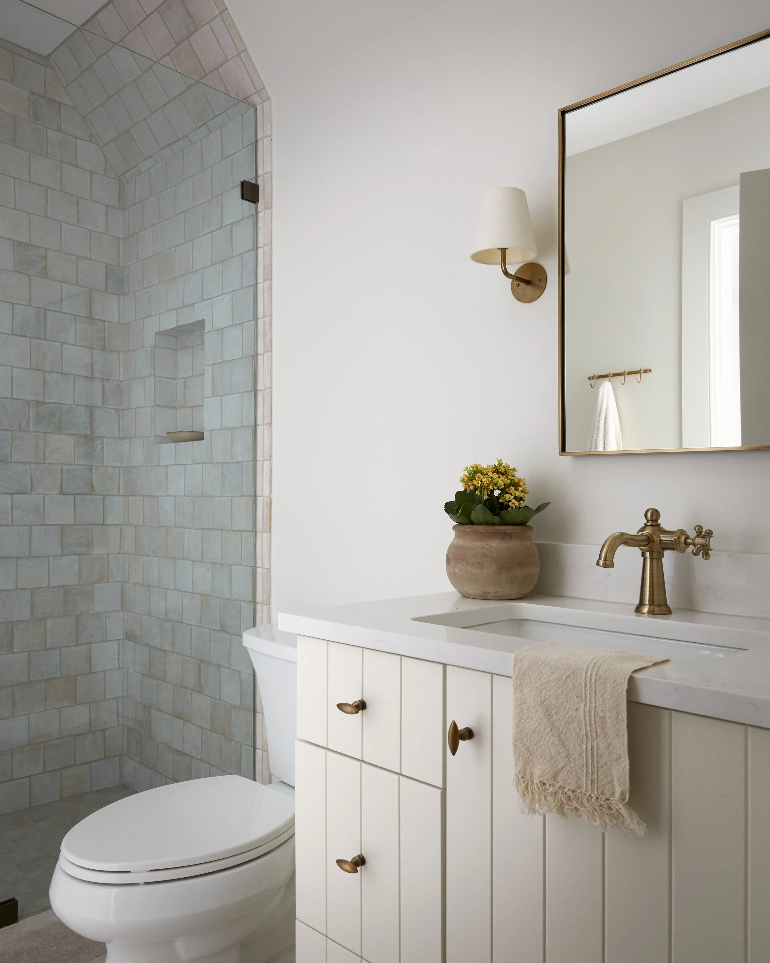 A bathroom with a white vanity, a large mirror, a brass faucet, a beige towel hanging on the vanity, a potted plant on the counter, a wall-mounted lamp, and a walk-in shower with beige tiles.