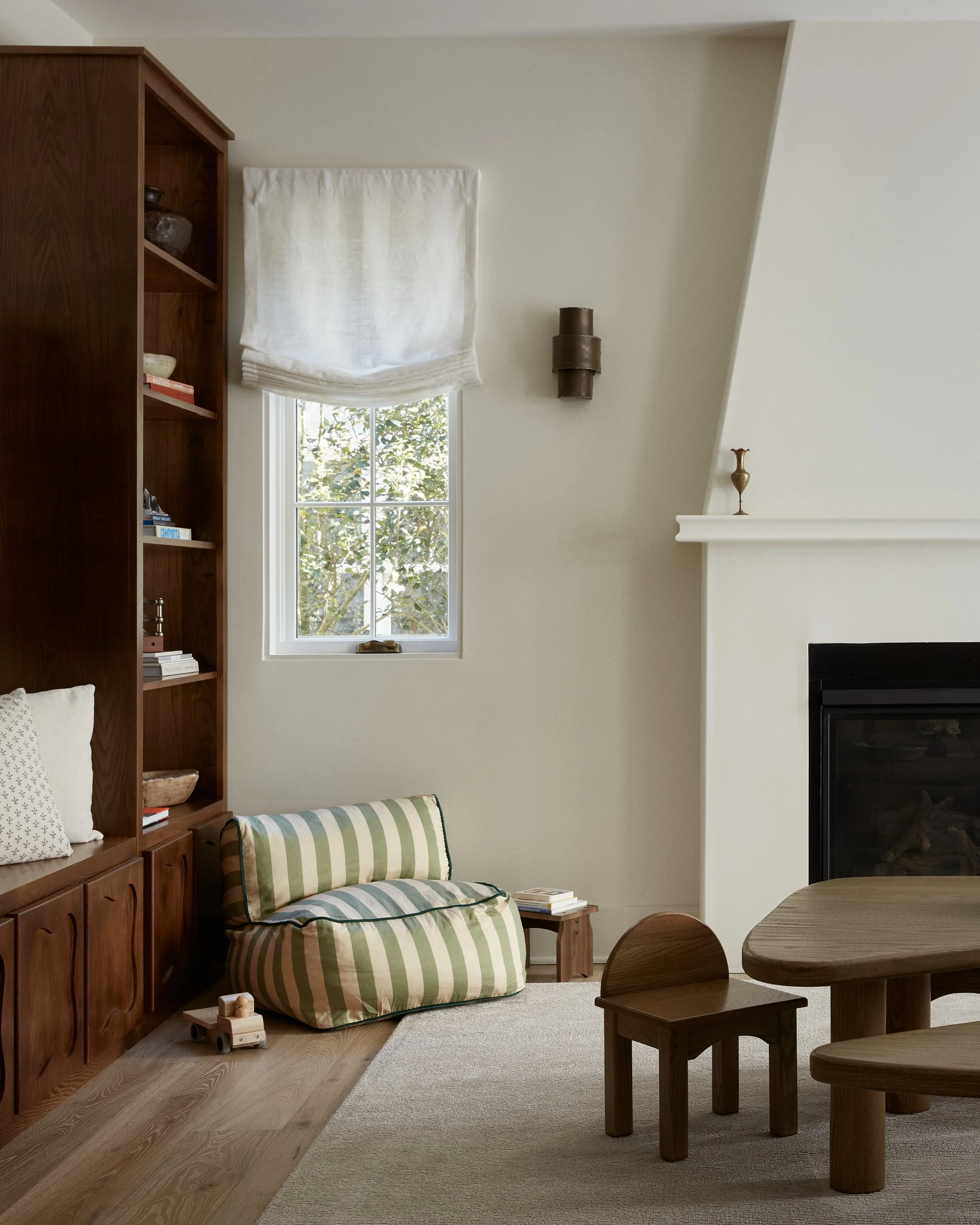 Living room with beige walls, a window with a white Roman shade, a wooden bookcase, a plaid loveseat, two wooden side tables, a round wooden dining table with chairs, a fireplace, and decorative items.