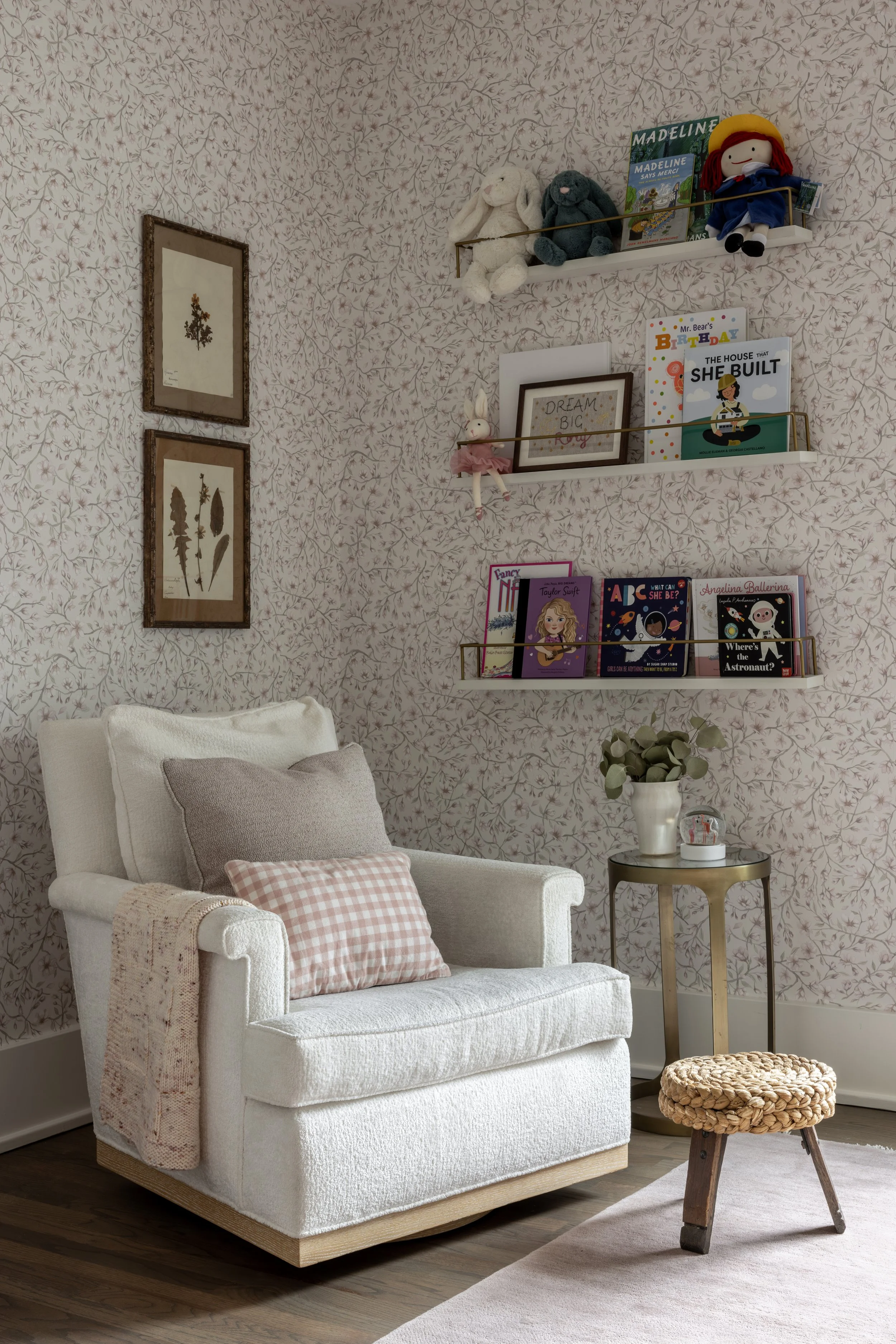 A cozy living room corner with a white upholstered armchair, beige and pink pillows, a small side table with a potted plant, a woven round ottoman, and wall shelves with children's books and plush toys, all against a floral wallpapered wall.