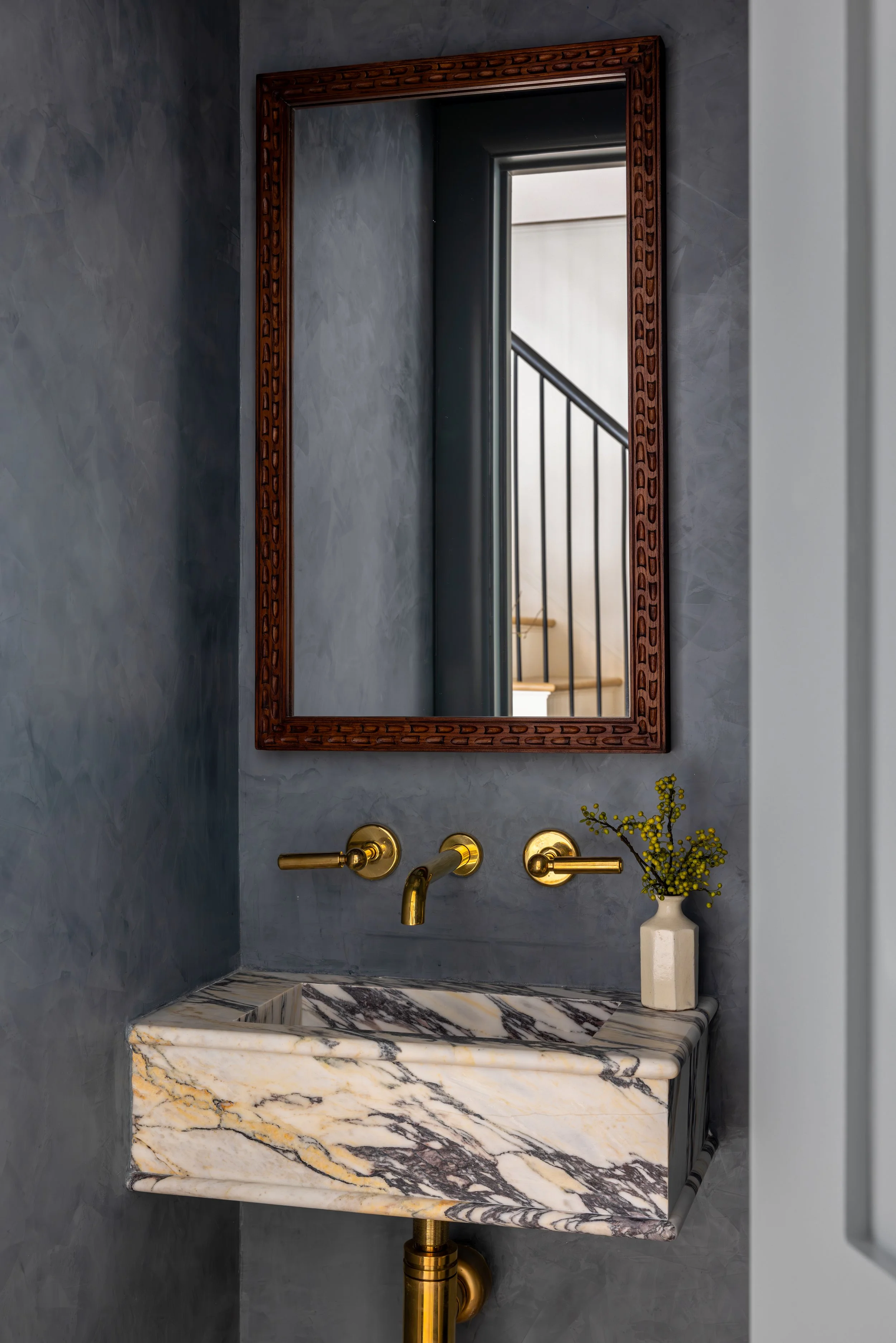 Modern bathroom sink with a marble countertop, a gold faucet, and a wall-mounted gold towel holder. A mirror with a wooden frame hangs above the sink, reflecting a staircase railing and window in the background. A small vase with yellow flowers is pl
