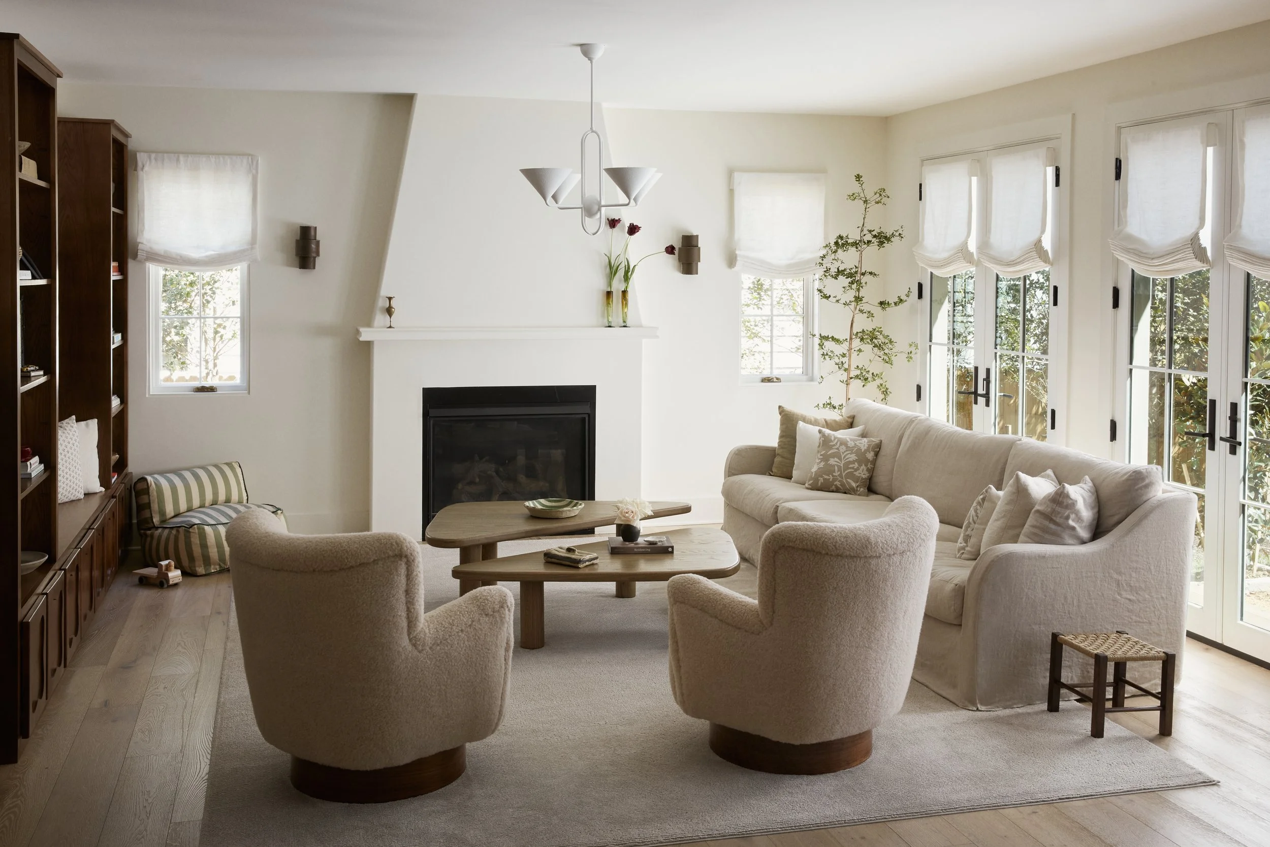 A cozy living room with a light beige sectional sofa, four armchairs, and a round wooden coffee table. Large windows with sheer white curtains allow natural light, and a white fireplace with floral decorations is on the wall. There is a built-in book