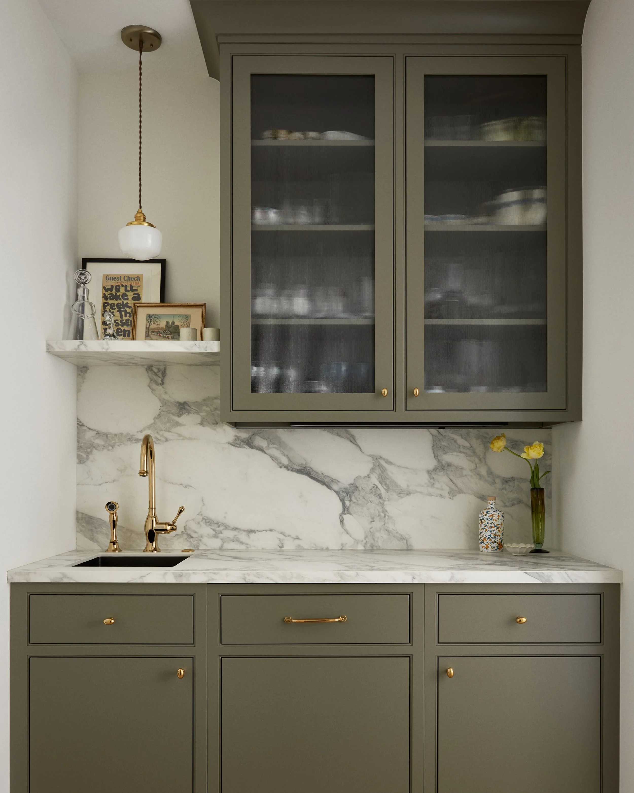 A kitchen corner with gray cabinets, a marble countertop, and a marble backsplash. There is a gold faucet, two vases with flowers on the right, and a small shelf with framed artwork, candles, and glassware on the left. A white pendant light hangs abo