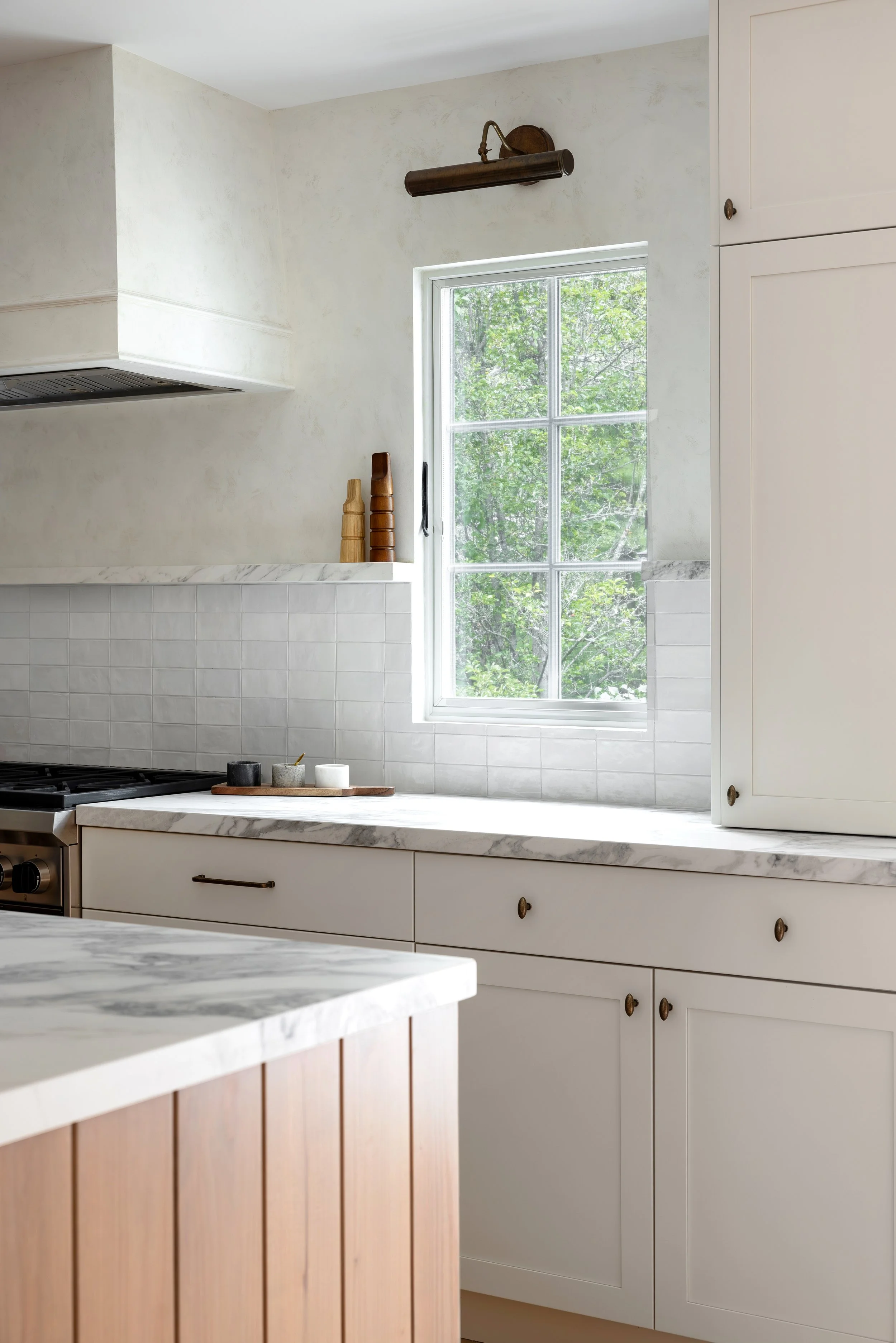Contemporary kitchen with white cabinetry, marble countertops, and a large window showing green trees outside.