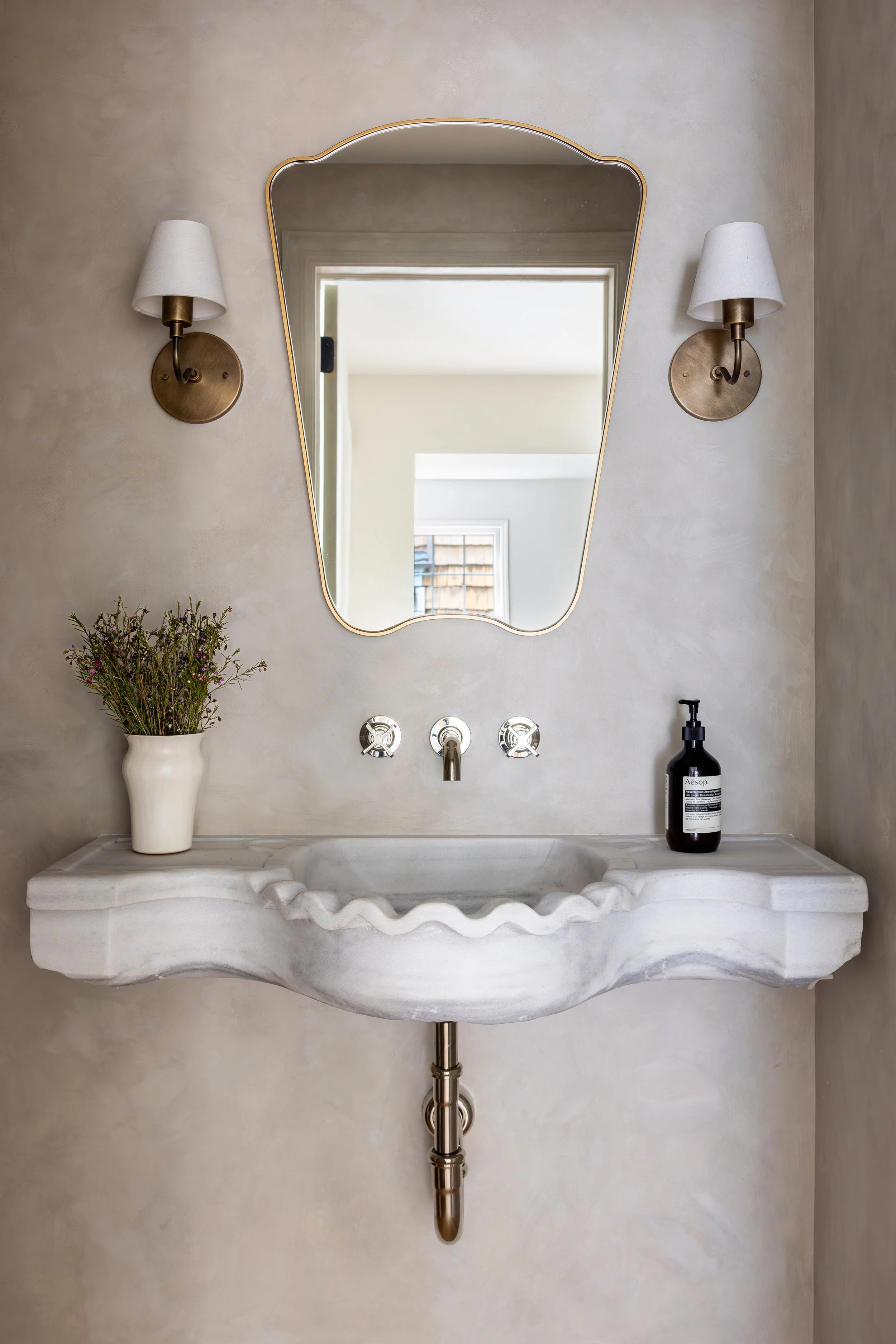 Bathroom vanity with a scalloped marble sink, a mirror, two wall sconces, a potted plant, and a soap dispenser.